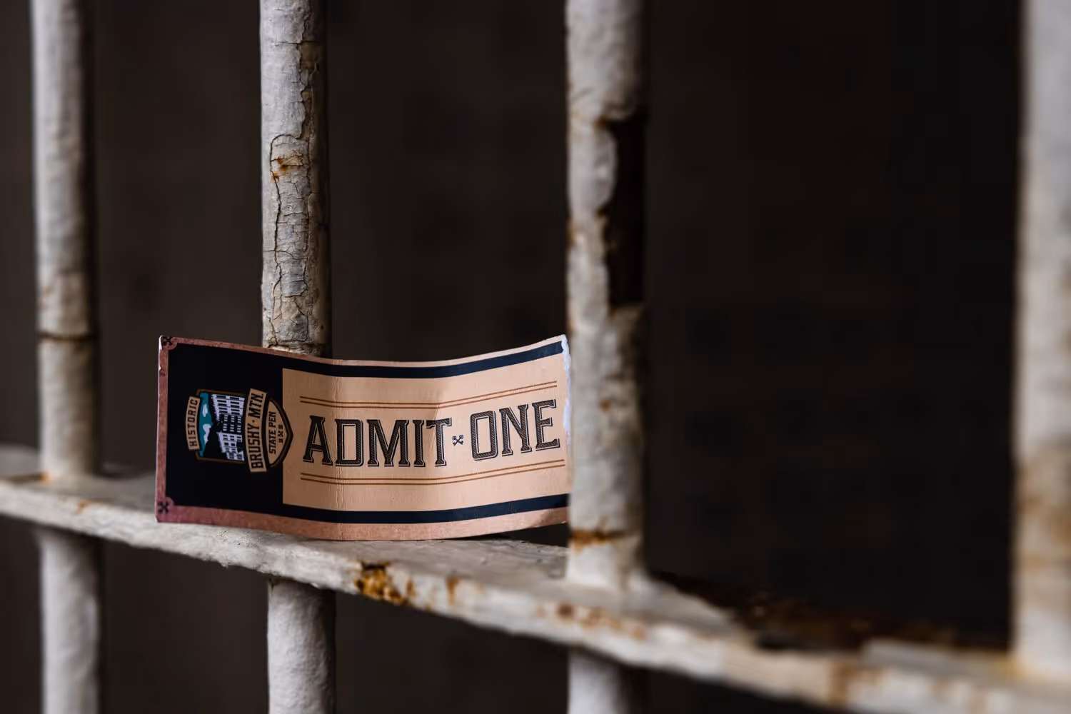 A Brushy admission ticket reading 'Admit One' placed on rusty prison bars.