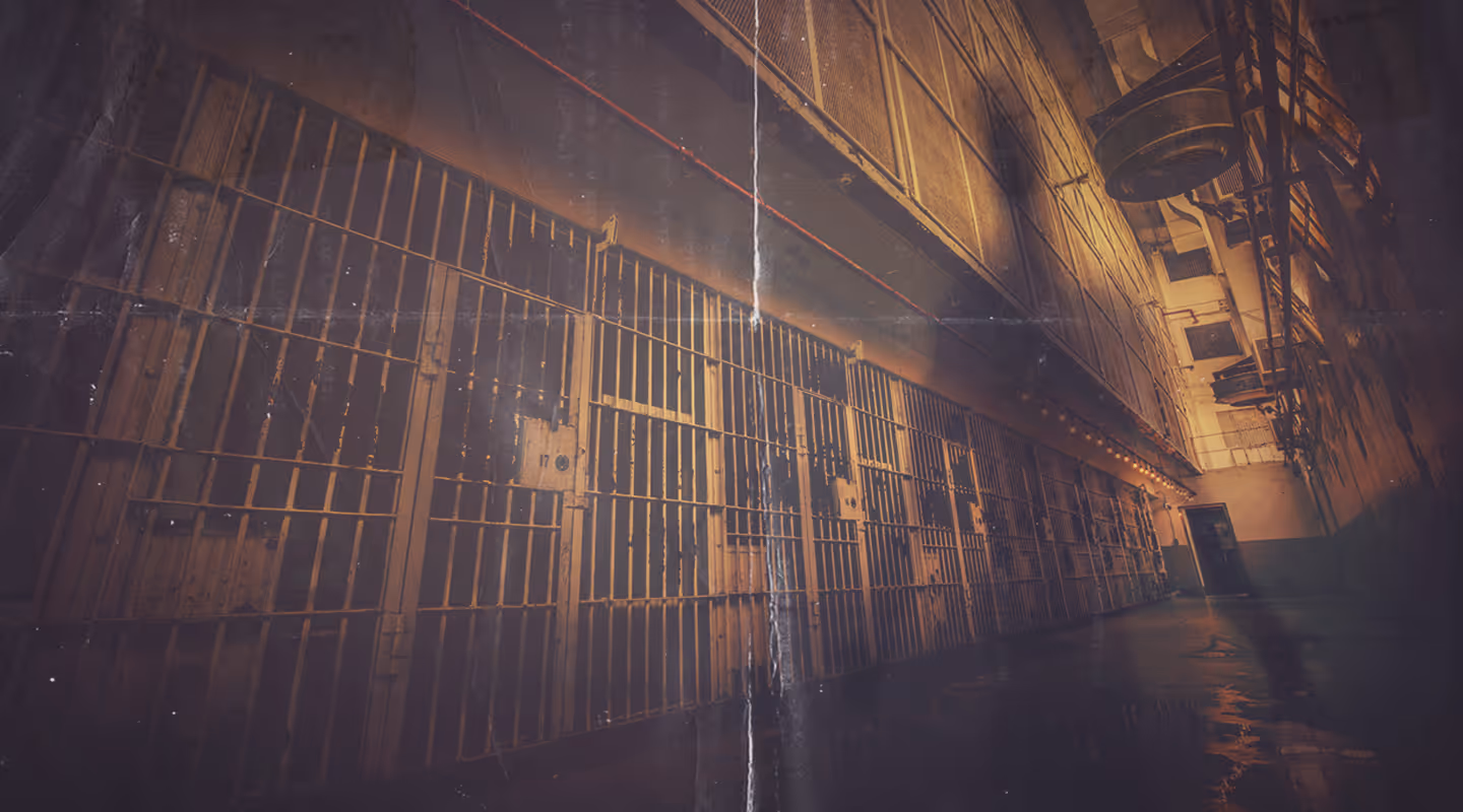 Dimly lit corridor of old prison cells with metal bars and a worn floor and a ghostly shadow silhouette of a person.