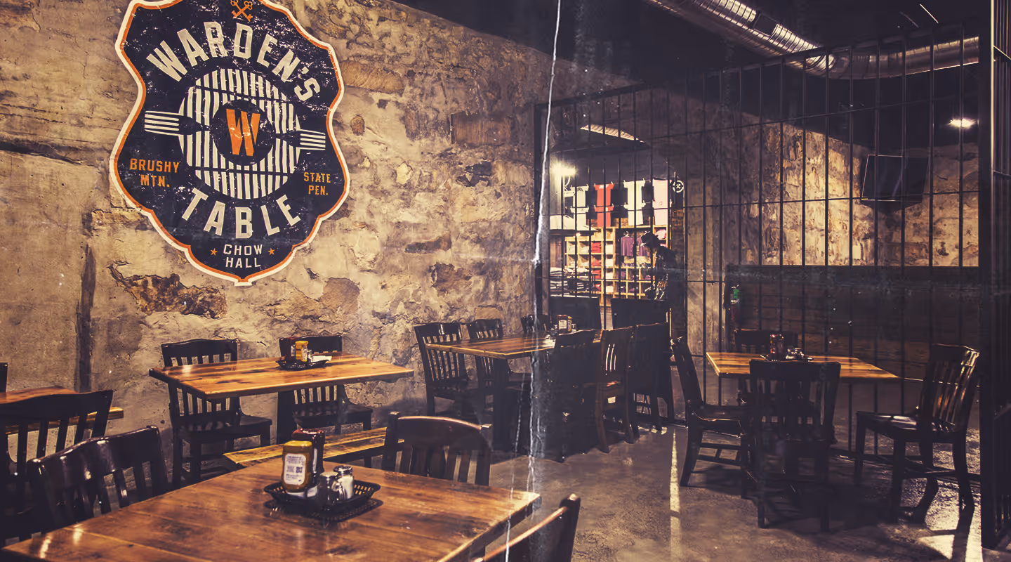 Rustic restaurant interior with wooden tables and chairs, stone walls, and a large Warden's Table logo sign on the wall.