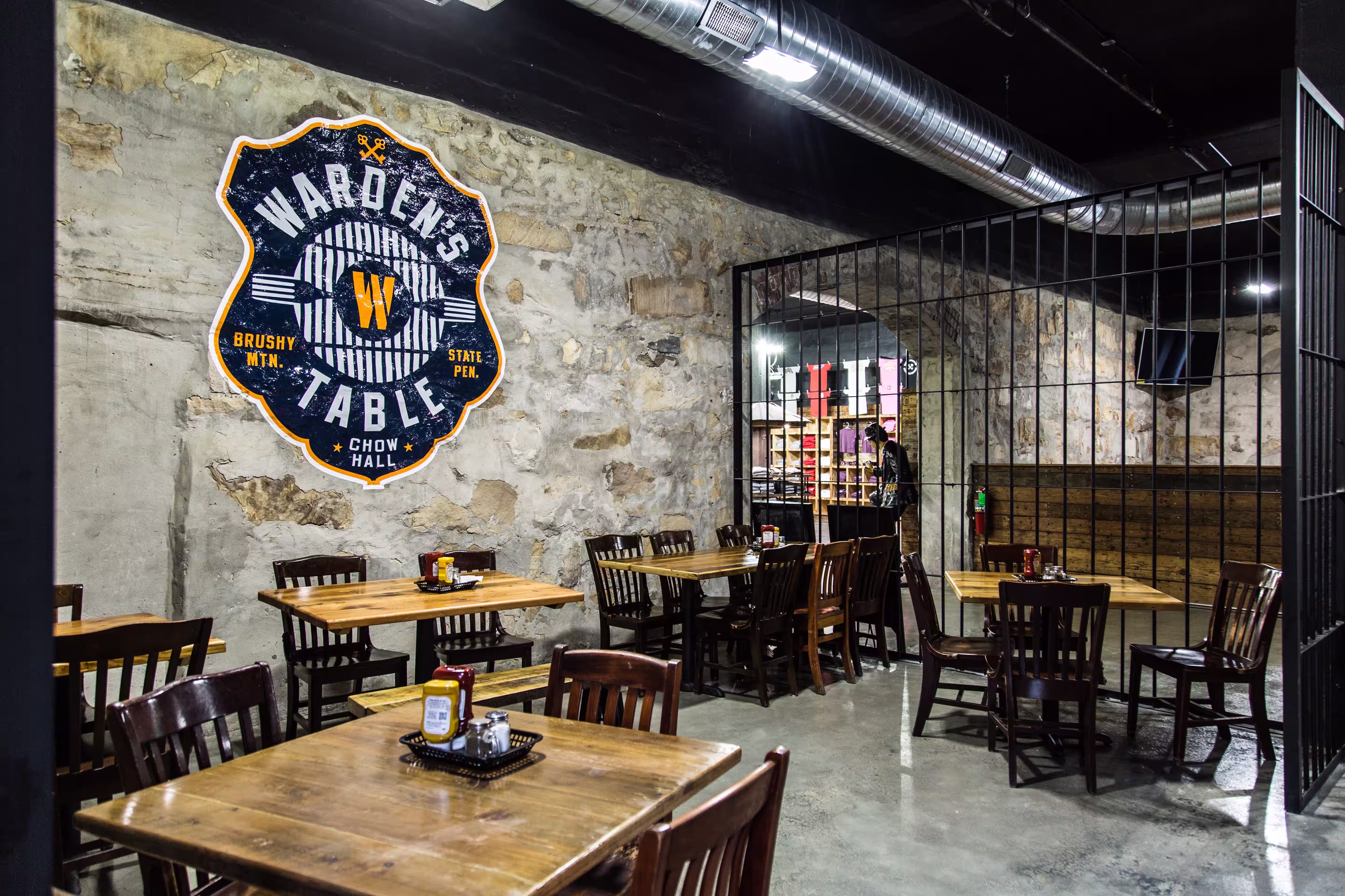 Interior of a rustic restaurant with wooden tables and chairs, stone walls, and a large Warden's Table sign on the wall.