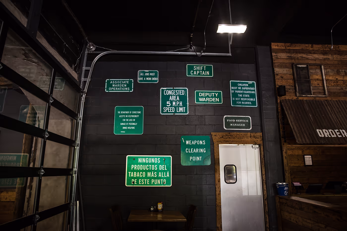 Restaurant wall with multiple green signs displaying various workplace and safety instructions above a small dining table and near a white door with window.