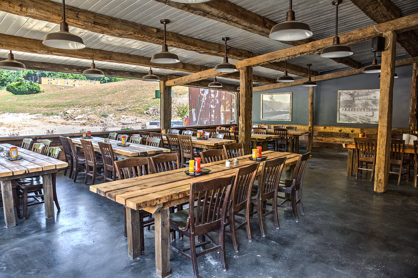 Rustic indoor dining area with wooden tables and chairs, condiment holders, hanging lights, and large windows showing a grassy hillside outside.