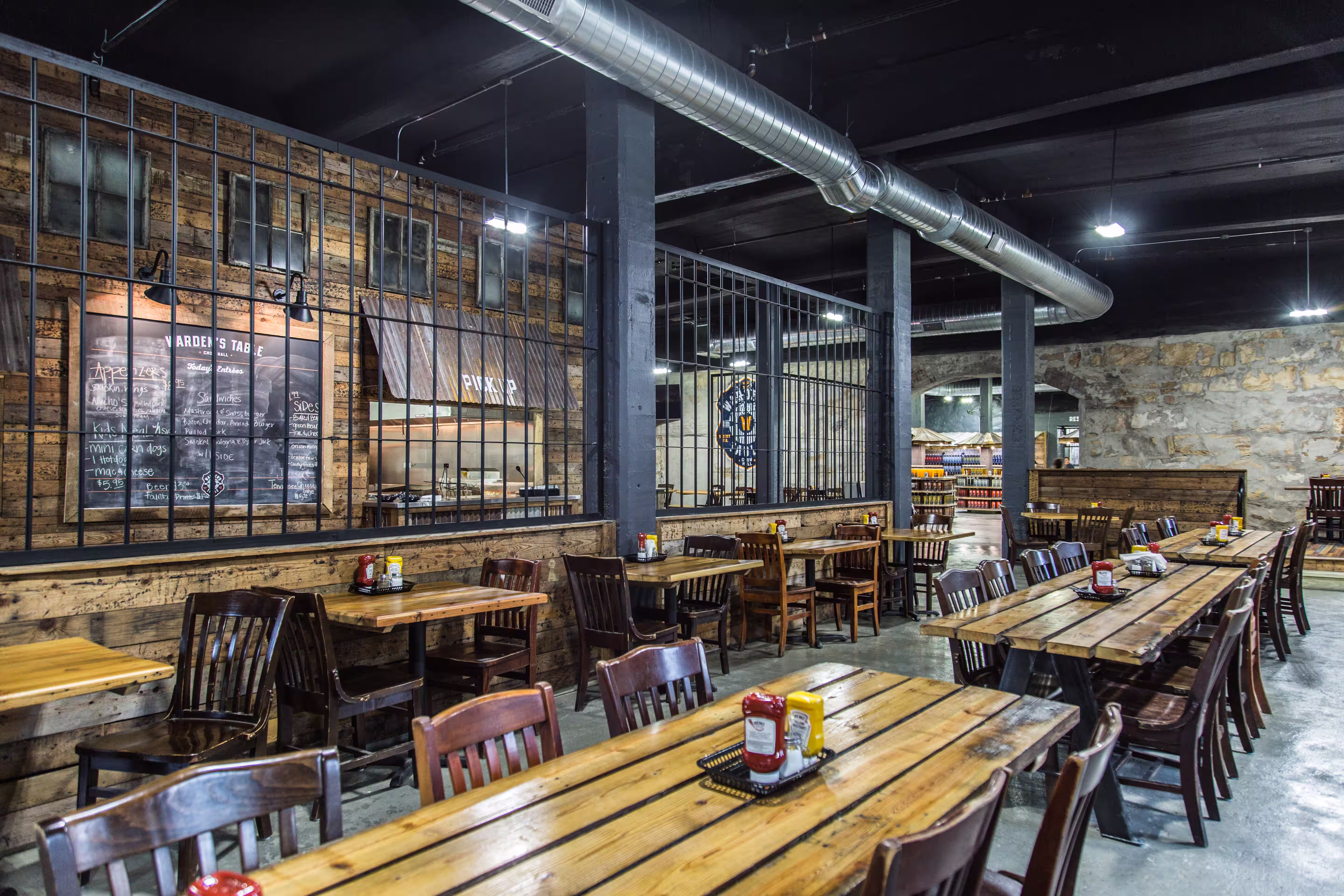 Rustic restaurant interior with wooden tables and chairs, condiment bottles on tables, industrial ceiling duct, and a menu board on the wall.