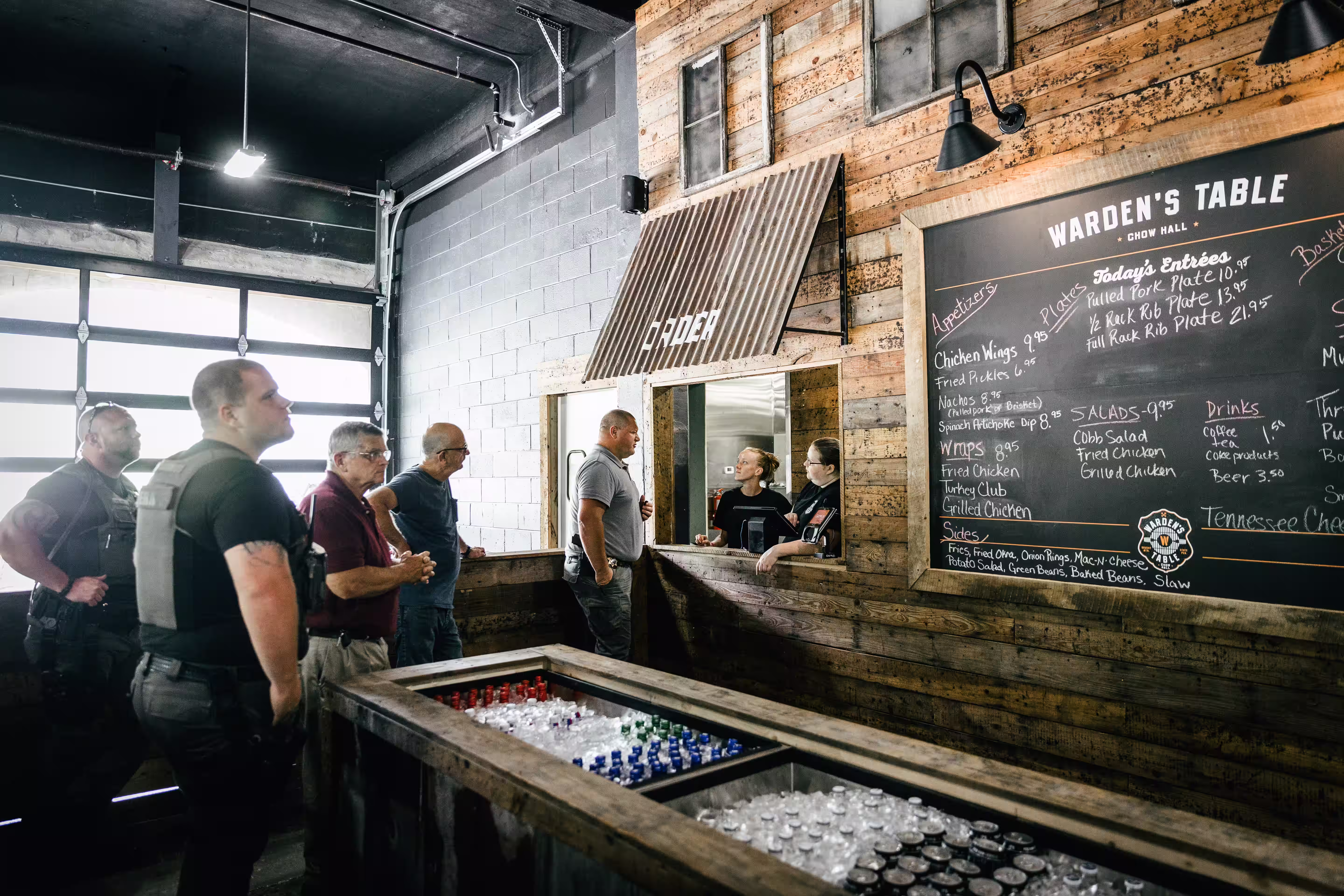 Customers and workers at Warden's Table restaurant ordering food at a rustic wooden counter with a large chalkboard menu on the wall.