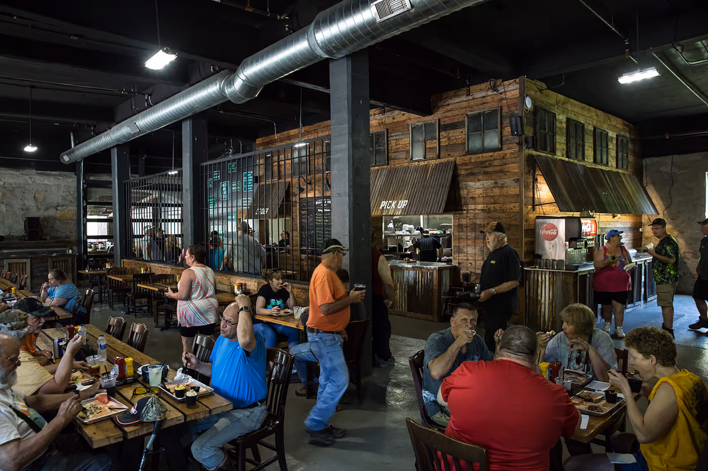 Busy rustic restaurant interior with wooden tables and walls, patrons eating and ordering at a pick-up counter.