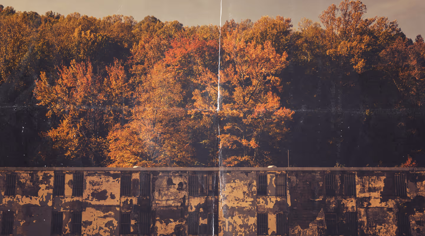 Old, weathered building with barred windows set against a forest of autumn trees with orange and yellow foliage.