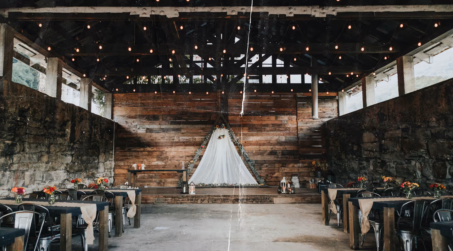 Rustic event space with stone walls, wooden stage featuring a triangular white drape with floral decorations, and tables adorned with burlap tablerunners and colorful flower centerpieces.