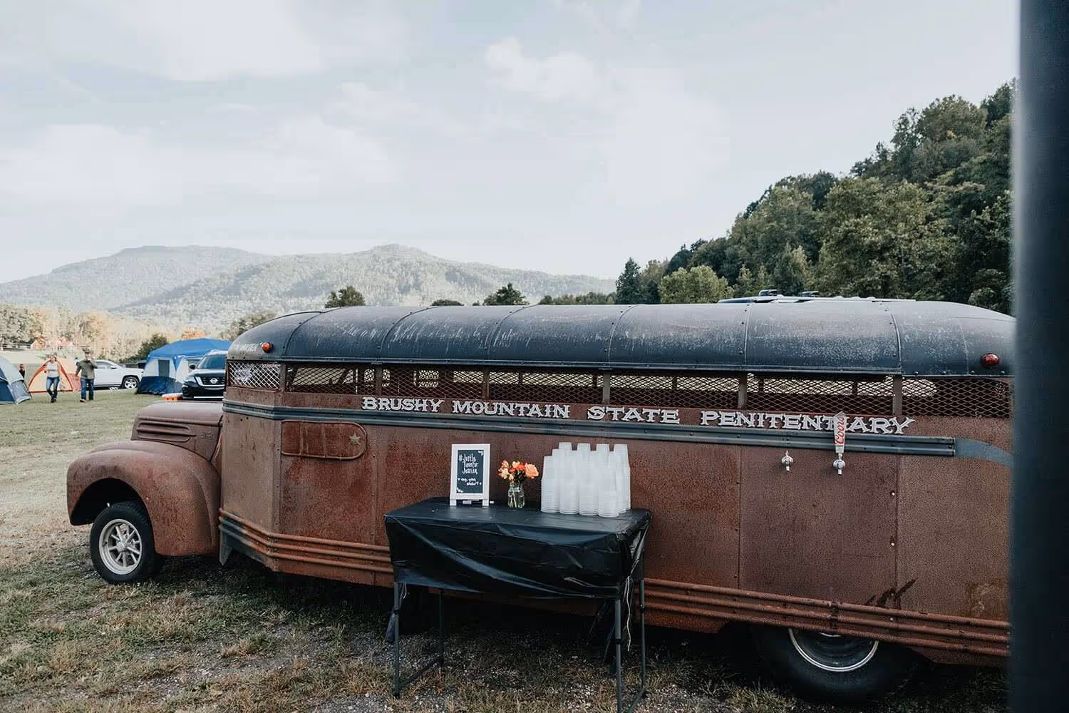 Rusty vintage bus labeled 'Brushy Mountain State Penitentiary' parked on grassy field with a table holding plastic cups and flowers nearby.