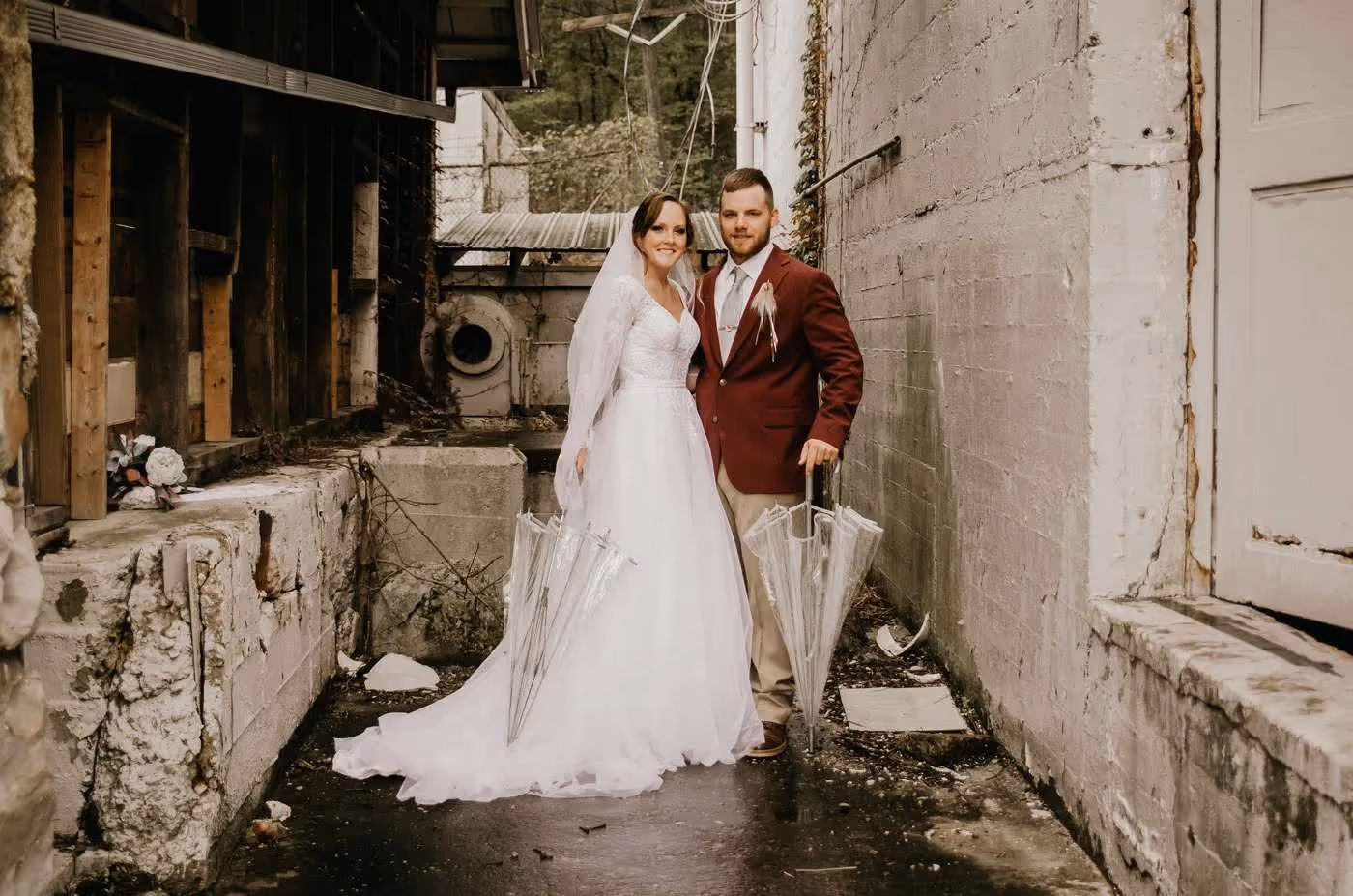 Bride in white wedding gown and groom in maroon jacket holding clear umbrellas in an alley with concrete walls.