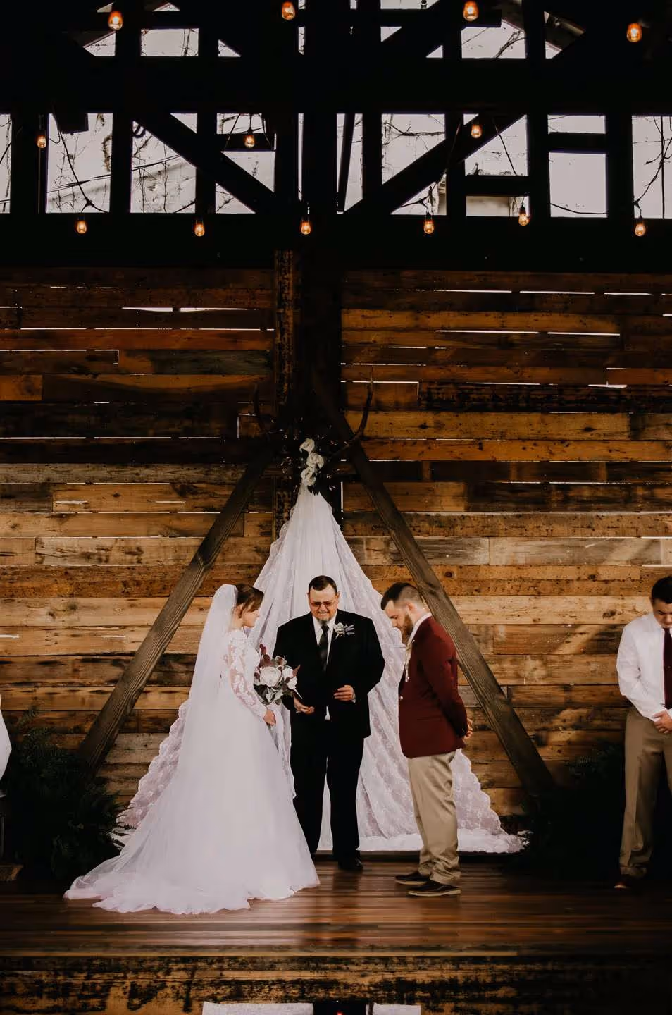 Bride and groom standing with an officiant in a rustic wooden venue decorated with string lights during a wedding ceremony.