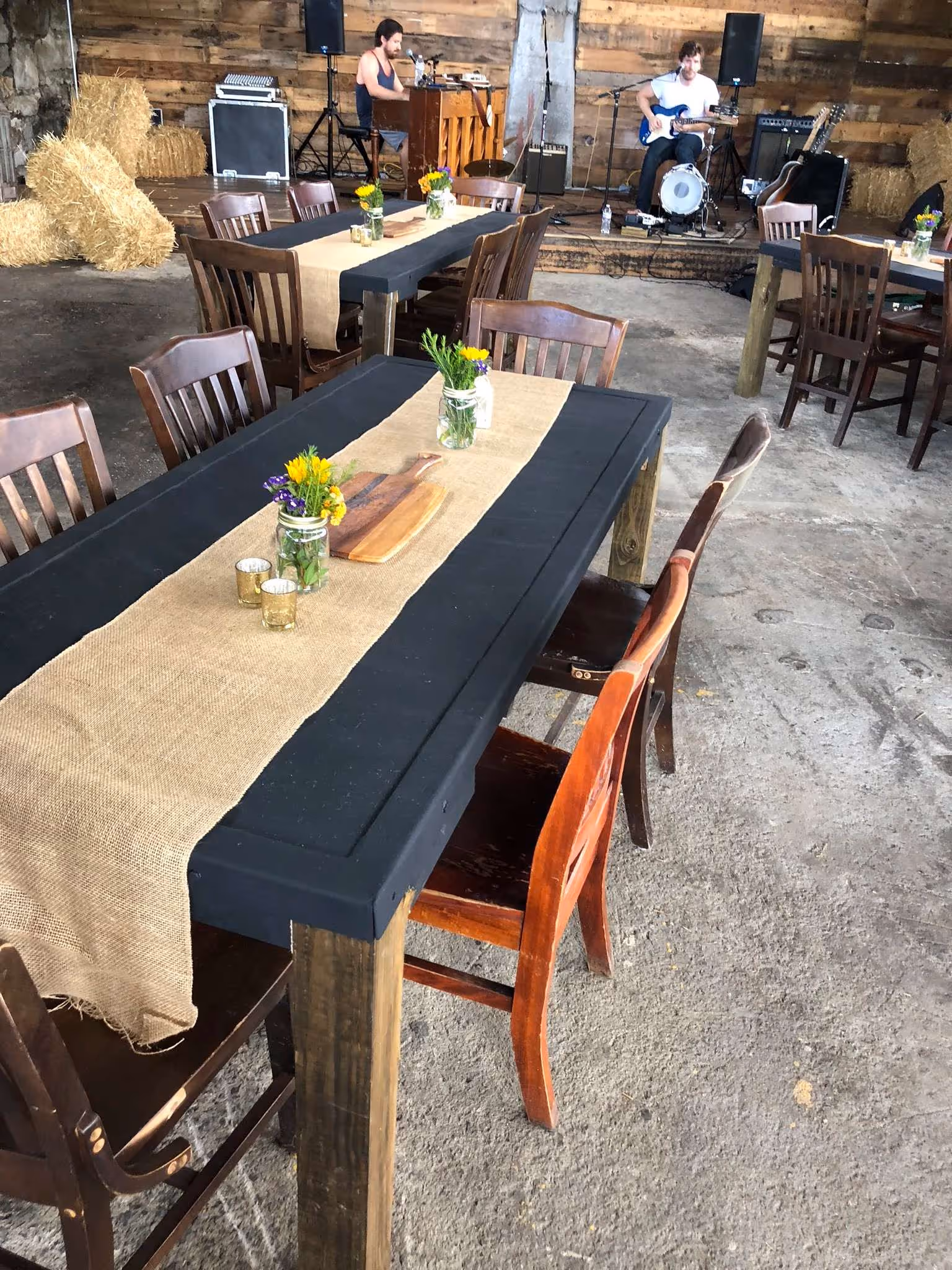 Rustic indoor event space with wooden tables and chairs, burlap runners, flower arrangements, hay bales, and musicians playing piano and guitar on a small stage.
