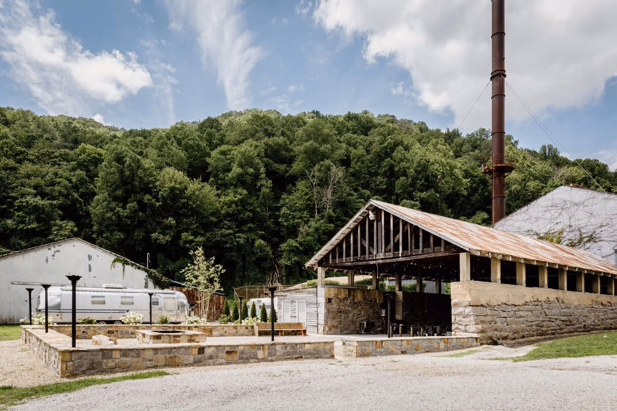 Outdoor scene of a rustic pavilion structure with stone walls and a rusty metal roof, set against a green forested hillside and blue sky with clouds.