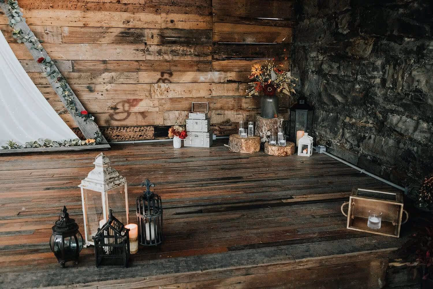 Rustic indoor scene with wooden floor and wall, decorated with lanterns, candles, flower arrangements, and stacked wooden boxes.