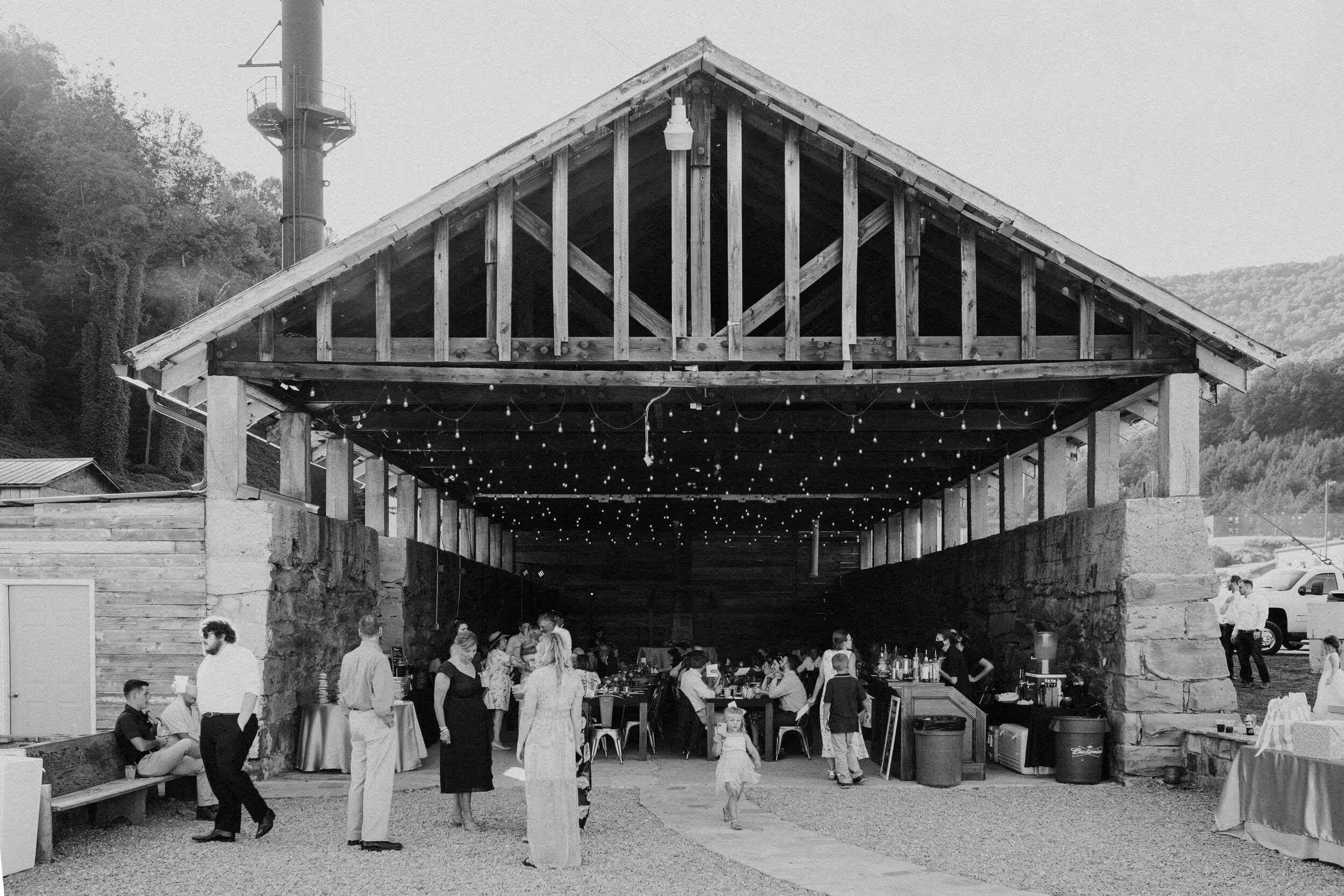 Black and white photo of people socializing inside and outside a rustic open wooden pavilion decorated with string lights.