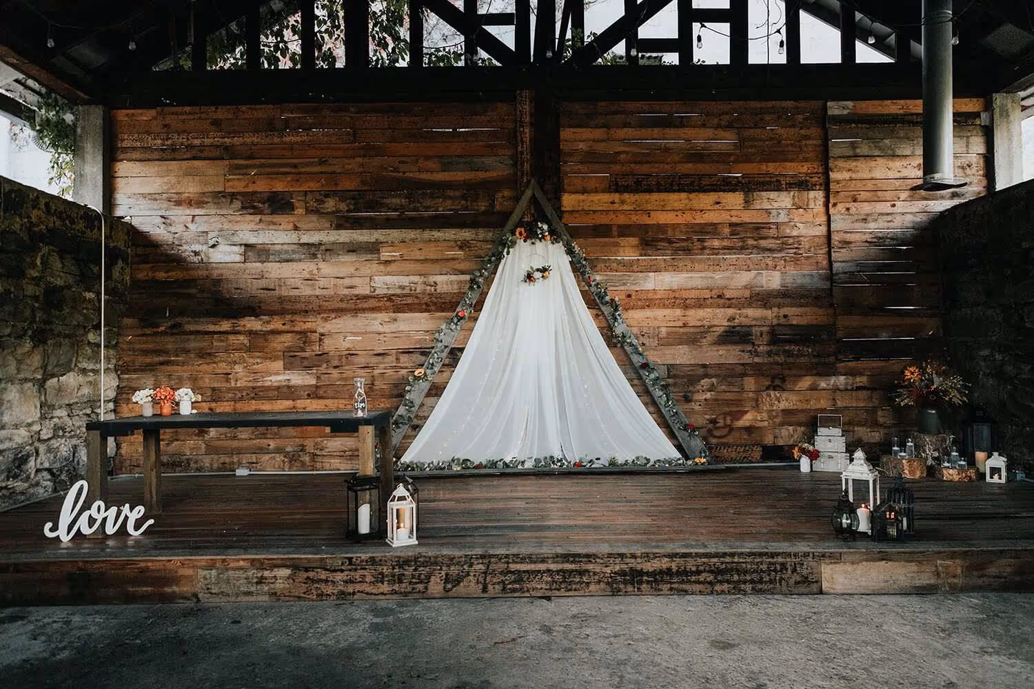 Rustic wedding altar with wooden backdrop, white fabric triangular frame decorated with greenery and flowers, small table, lanterns, and a 'love' sign.