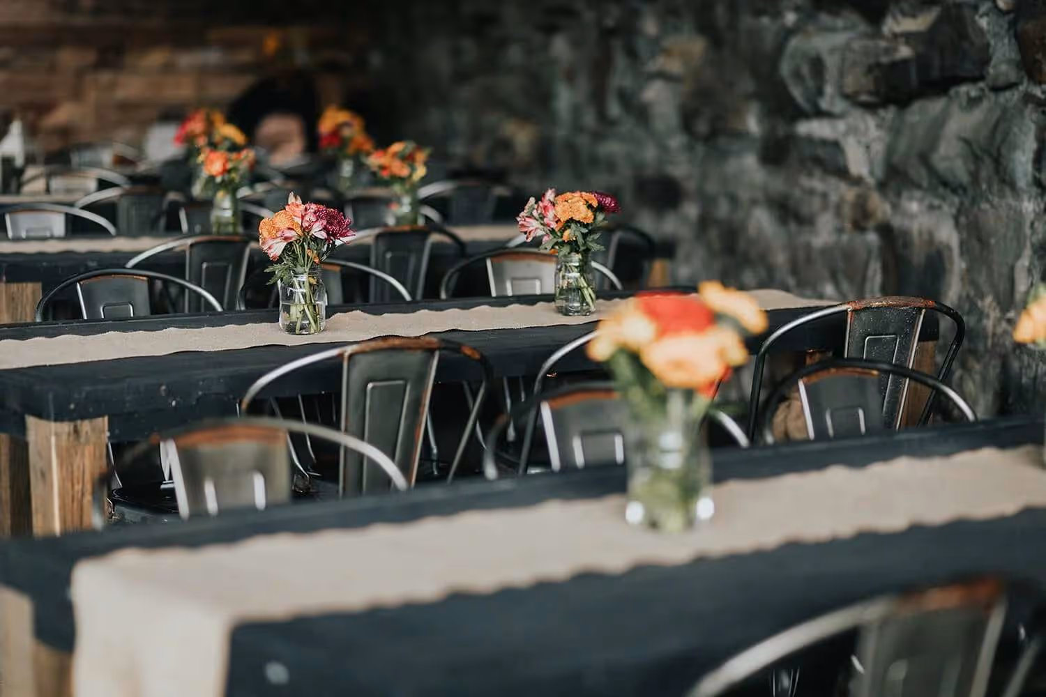 Rustic tables with burlap tablecloth runners and glass jars holding colorful flower arrangements in a dimly lit stone-walled room.