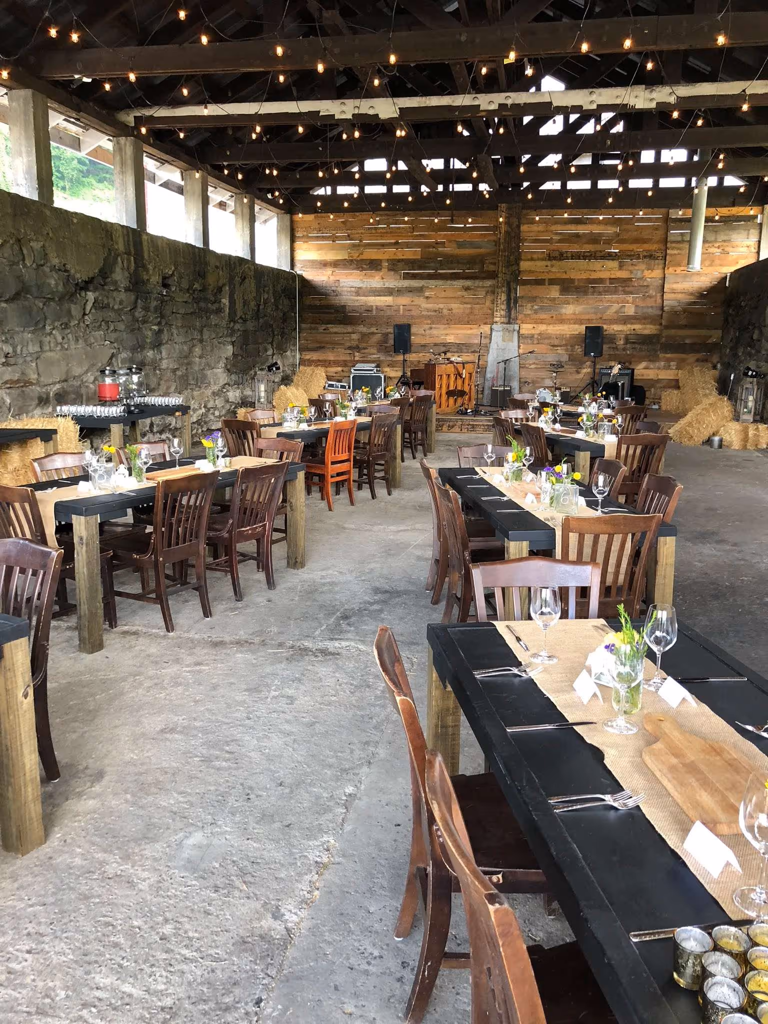 Rustic event space with long wooden tables set with glasses, cutlery, and small flower arrangements, string lights overhead, and hay bales along stone walls.
