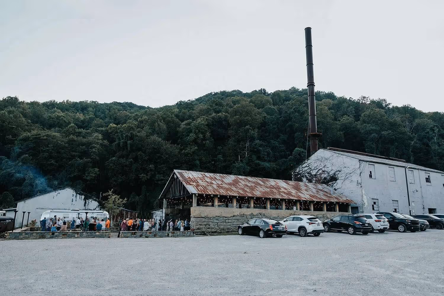 Rustic venue with white buildings and a tall chimney surrounded by forest, with people gathered near an Airstream trailer and cars parked in the gravel lot.