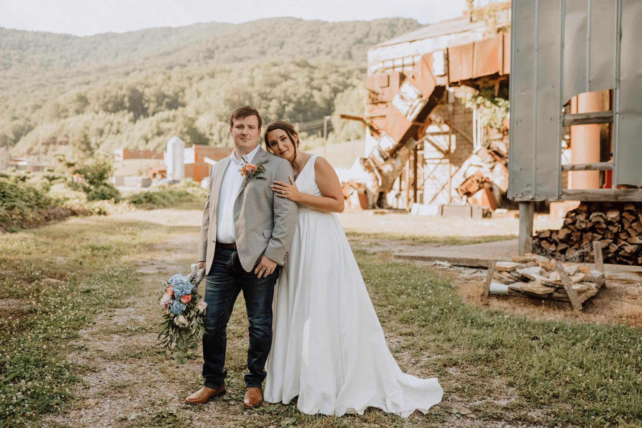 Bride in white dress leaning on groom in light gray jacket holding a bouquet outdoors with greenery and rustic structures in the background.