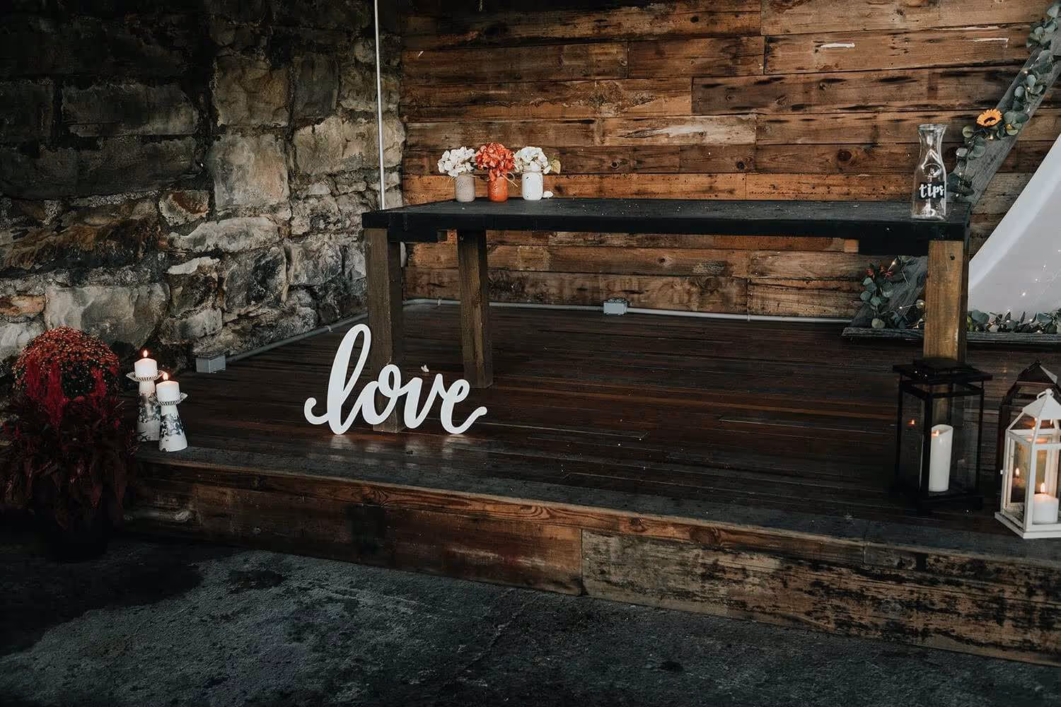 Rustic wooden stage with white cursive 'love' sign, candles, flowers, and lanterns.