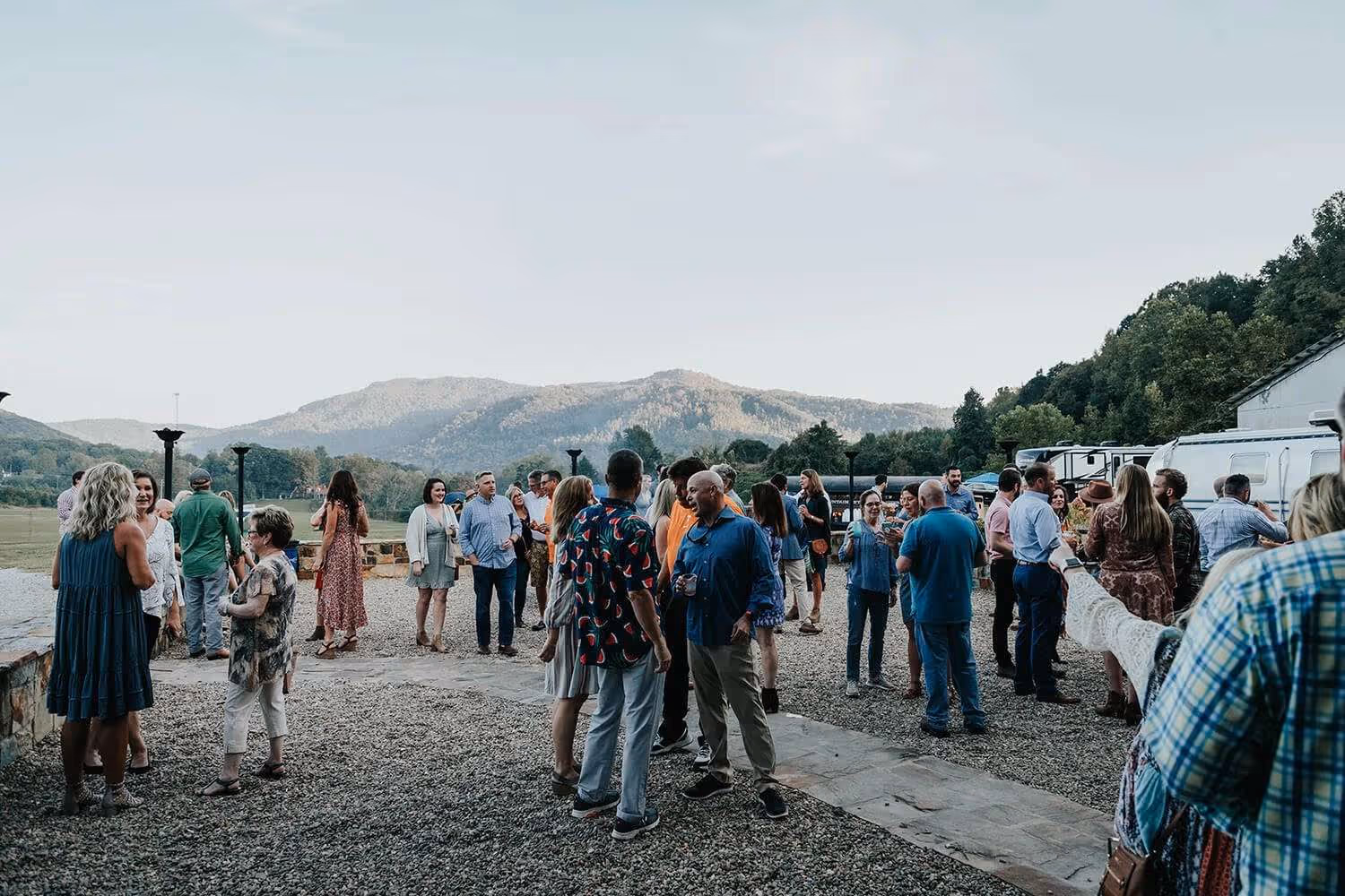 A group of people socializing outdoors on a gravel area with mountains and trees in the background under a clear sky.