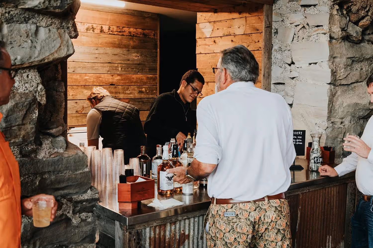 Two bartenders serving drinks to men at a rustic bar with stone walls and wooden panels.