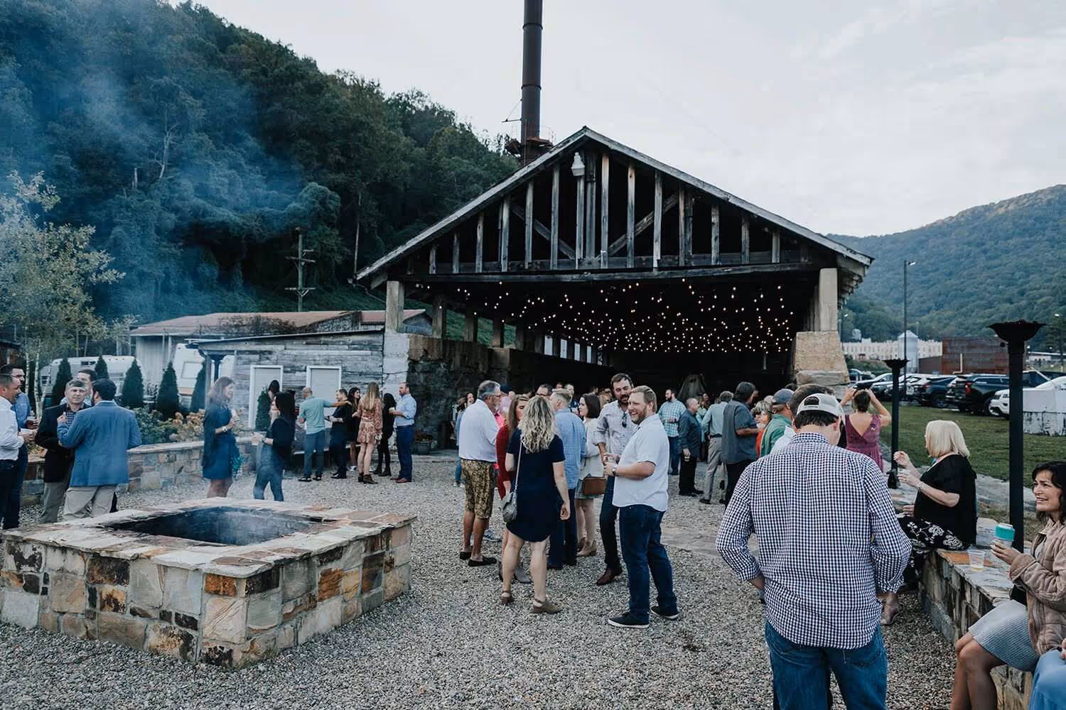 Outdoor gathering with people socializing near a stone fire pit and a rustic pavilion decorated with string lights in a mountainous setting.