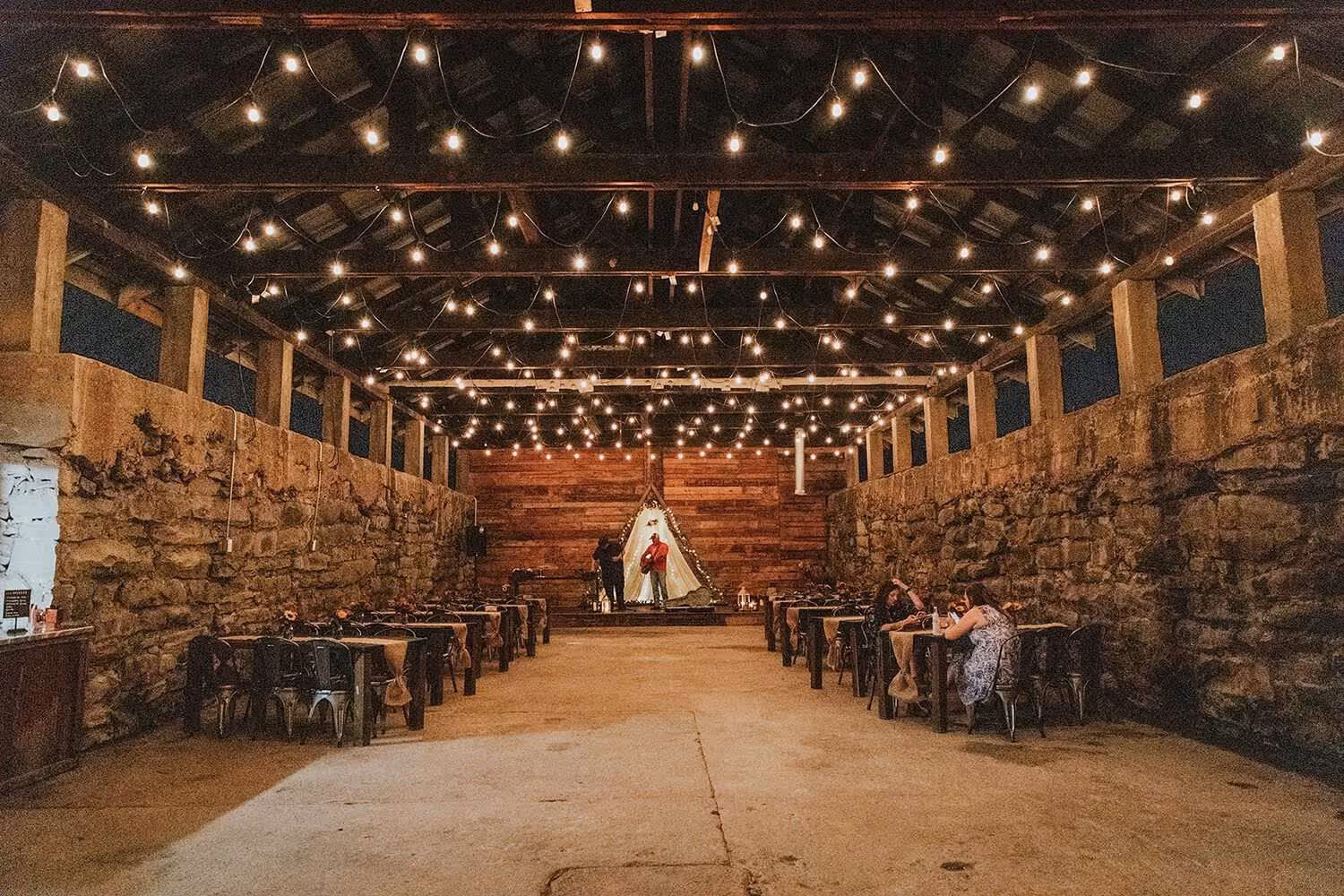 Rustic event hall with stone walls, string lights overhead, tables with chairs on both sides, and two people near a small illuminated stage.