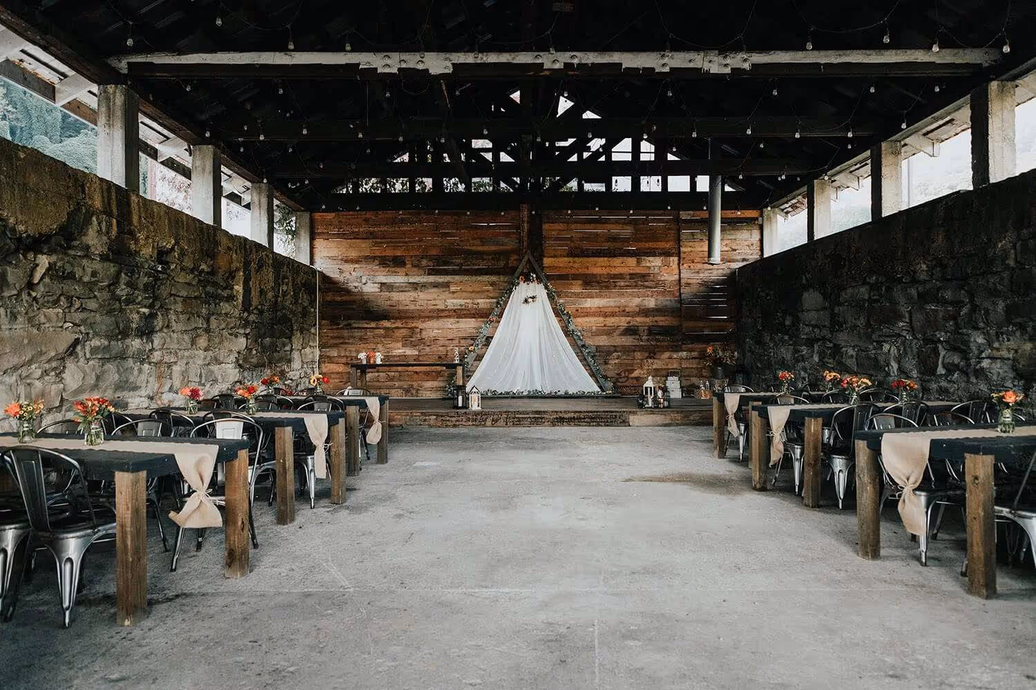 Rustic wedding reception area with wooden tables, metal chairs, and floral centerpieces under a wooden roof with stone walls.