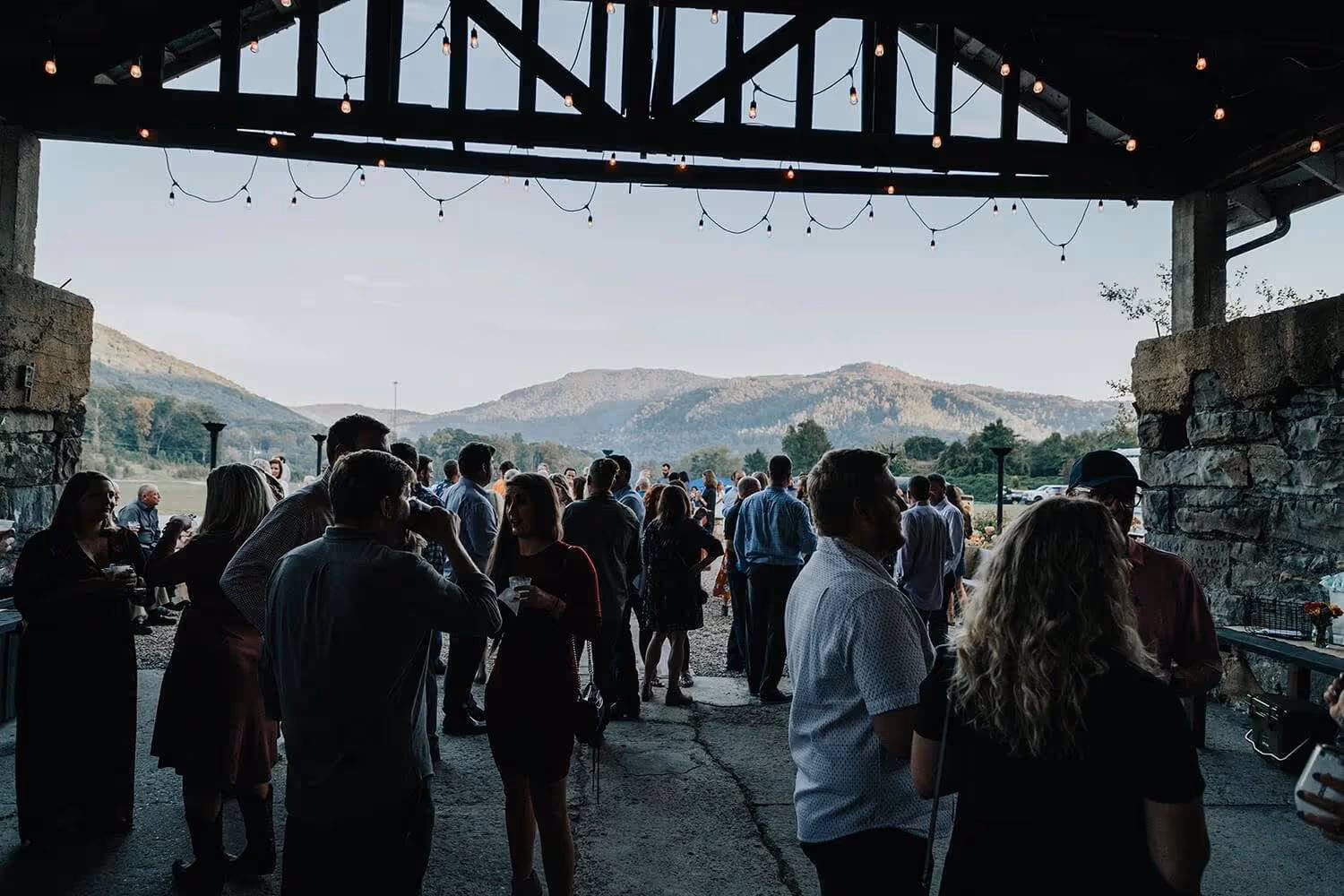 Group of people socializing under a covered pavilion with hanging string lights and mountains in the background.
