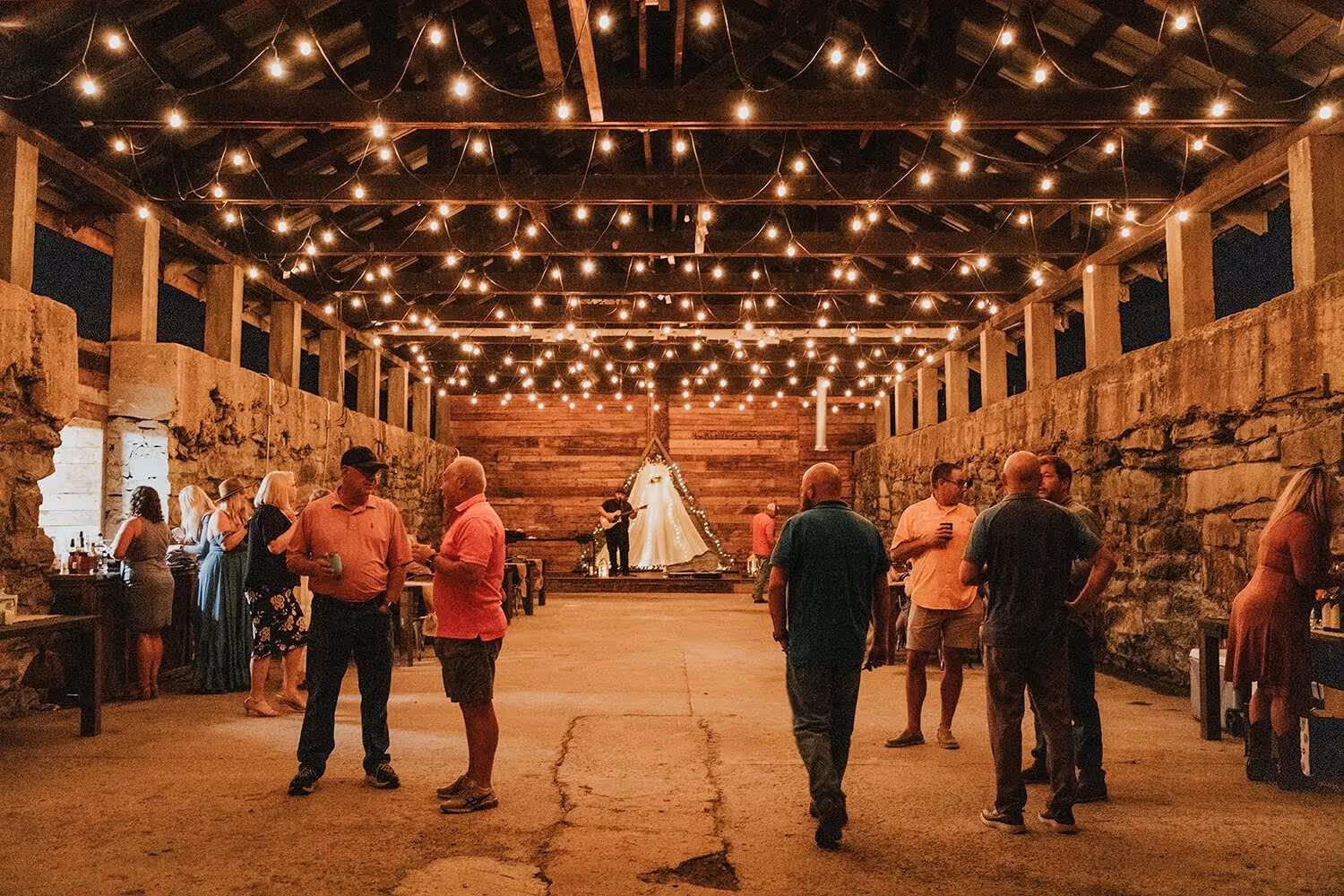 People socializing under string lights inside a rustic stone and wood pavilion with a musician playing guitar on a lit stage in the background.