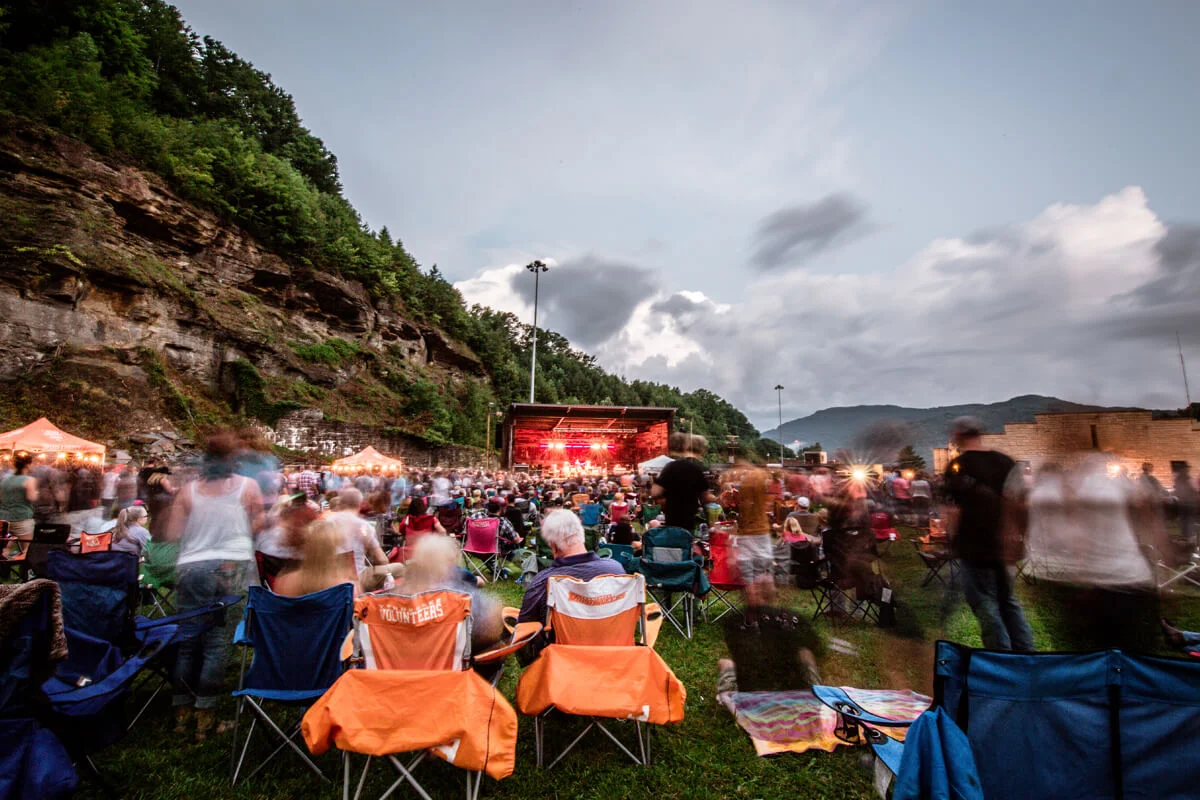Outdoor Jamey Johnson concert with a crowd sitting on chairs and blankets on grass, surrounded by rocky cliffs and trees at dusk.