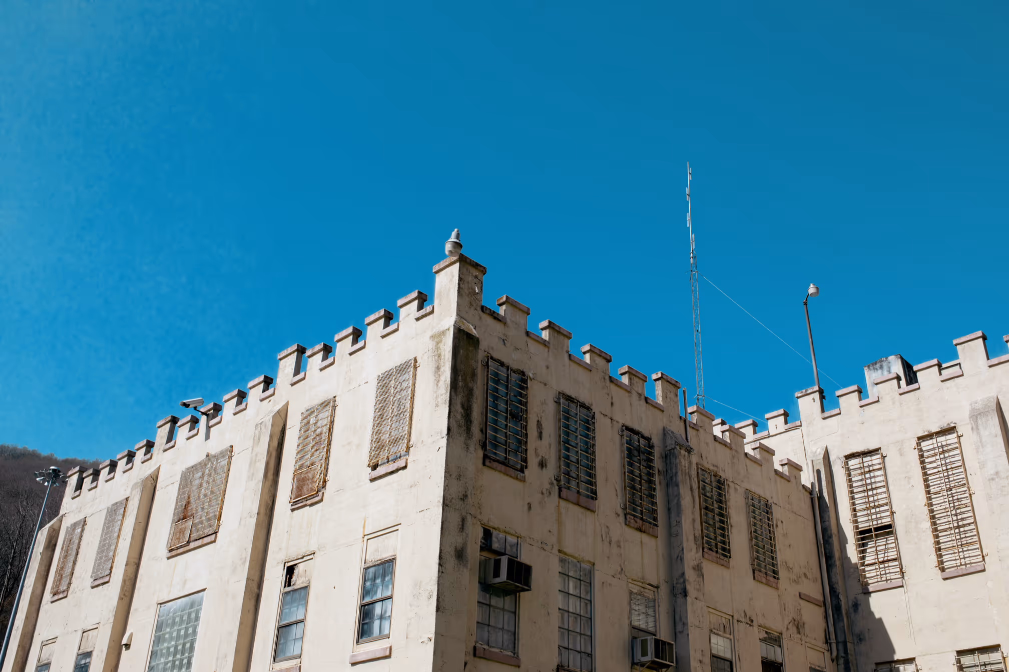 Exterior corner view of a beige prison building with barred windows, security cameras, and a tall antenna under a clear blue sky.