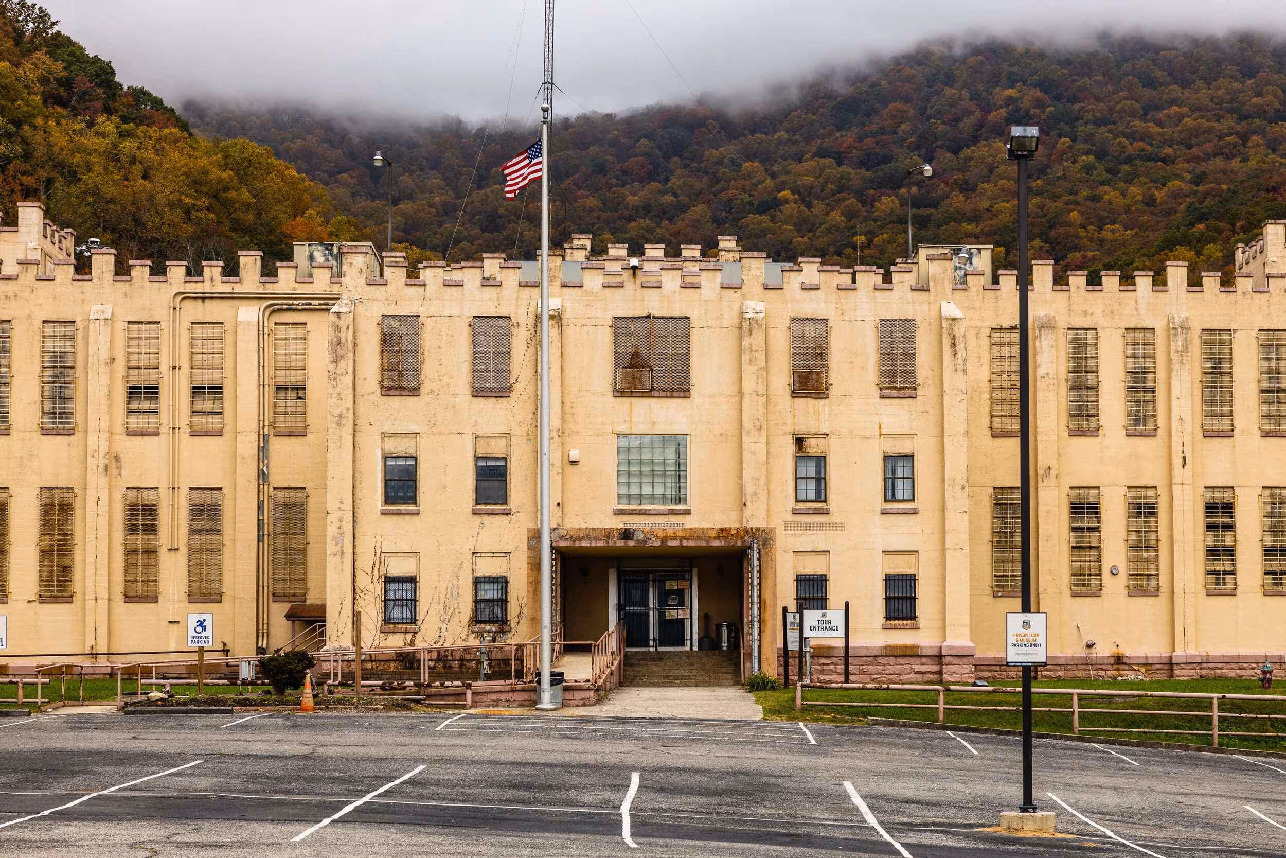 Front view of a beige prison building with barred windows, an American flag at half-mast, and a cloudy mountain backdrop.