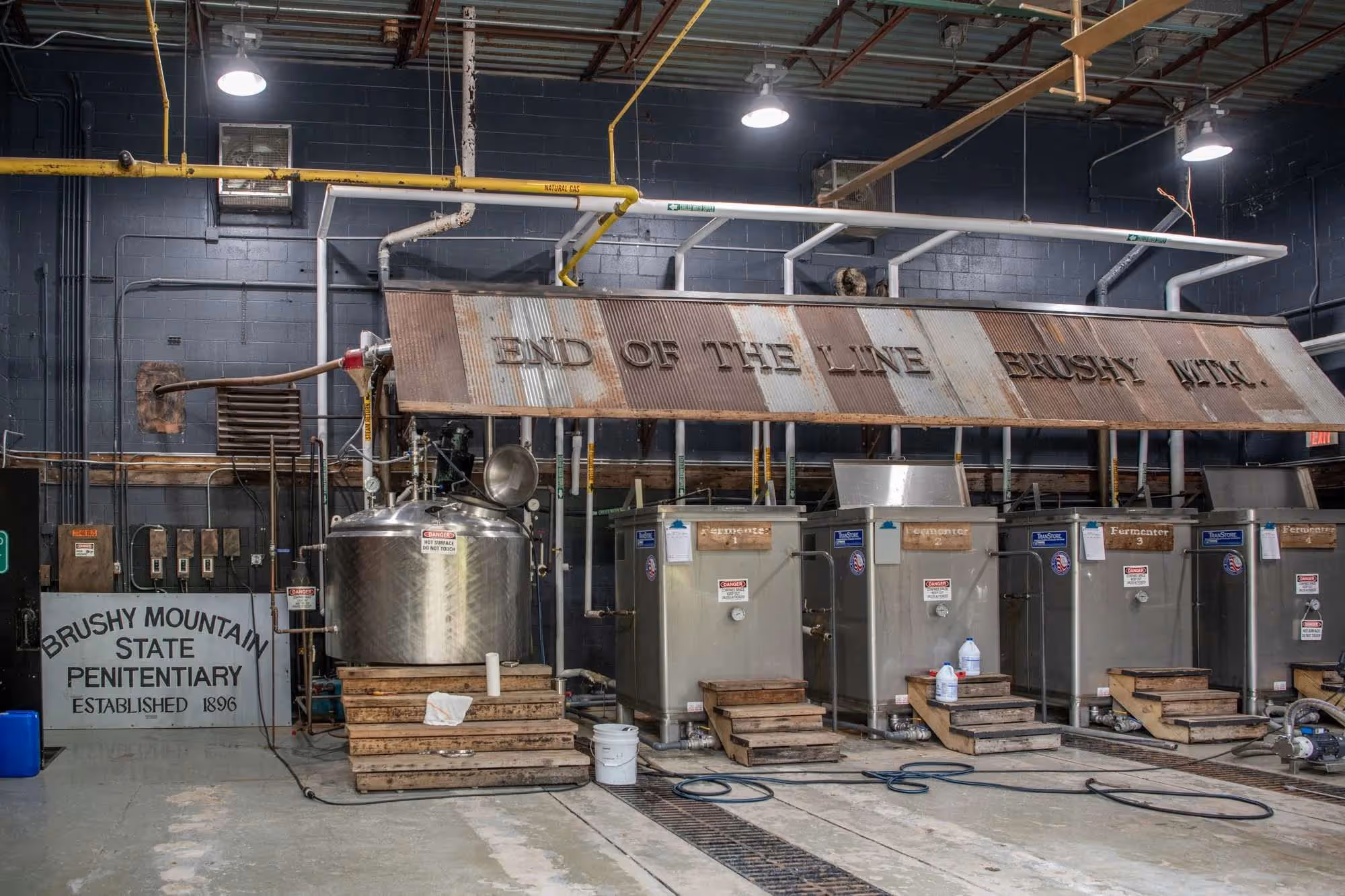 Interior view of Brushy Mountain State Penitentiary Distillery with metal fermenter tanks labeled 1 to 4 and large rusty sign reading 'END OF THE LINE BRUSHY MTN.'