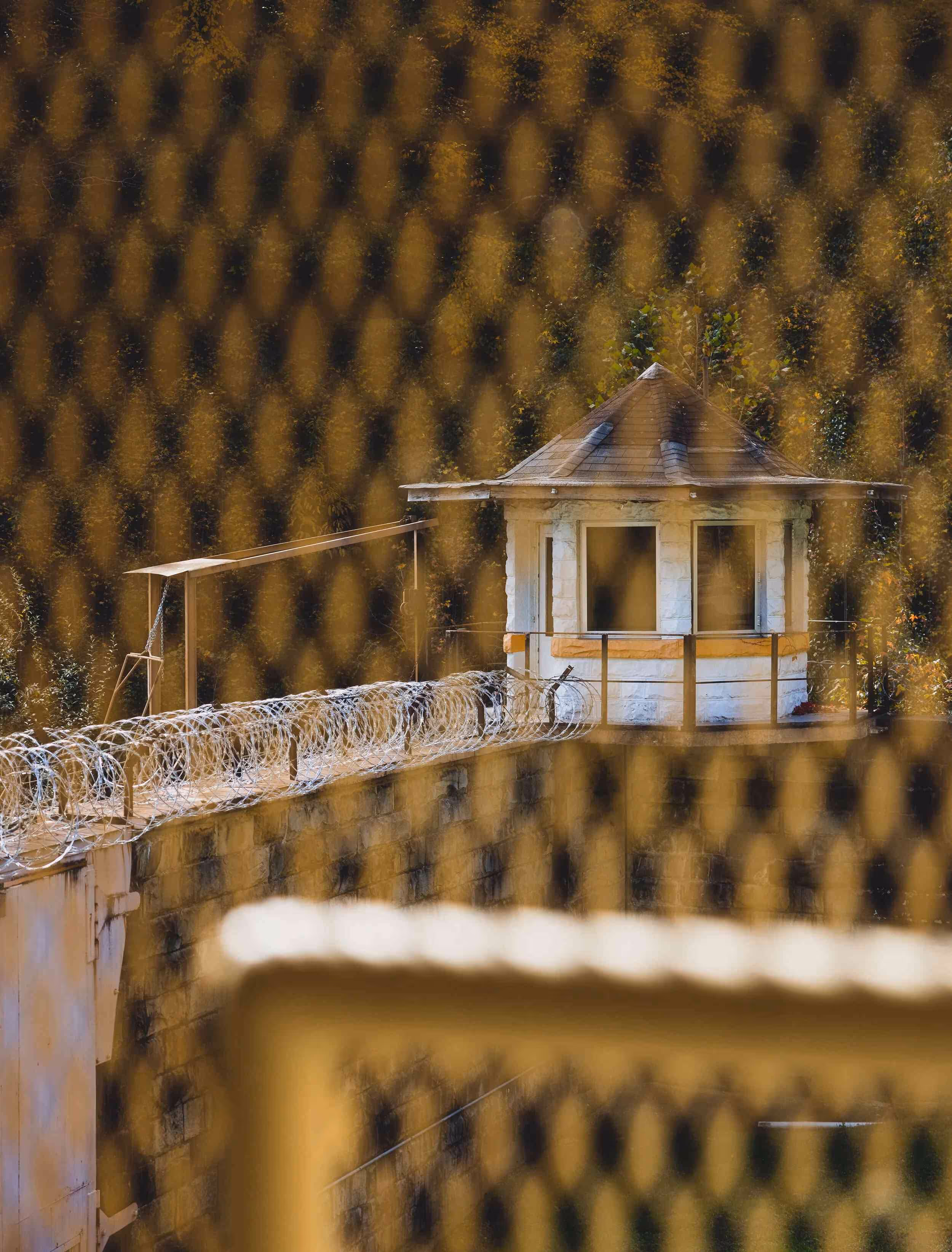 View through a mesh fence of a white guard tower with a barbed wire fence on a stone wall surrounded by trees.