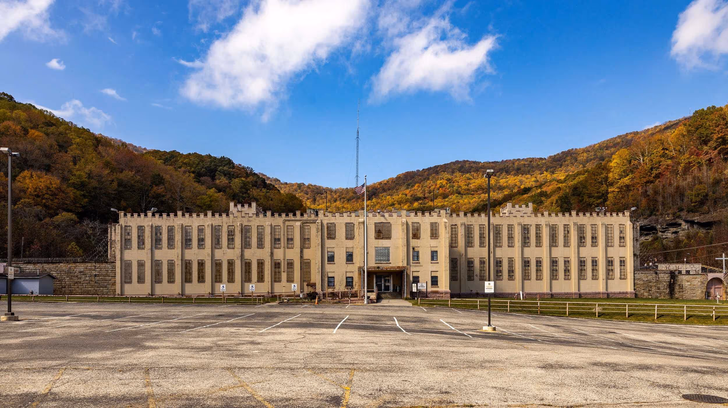 Large rectangular beige prison building with barred windows and an American flag outside, surrounded by forested hills with autumn foliage under a partly cloudy blue sky.