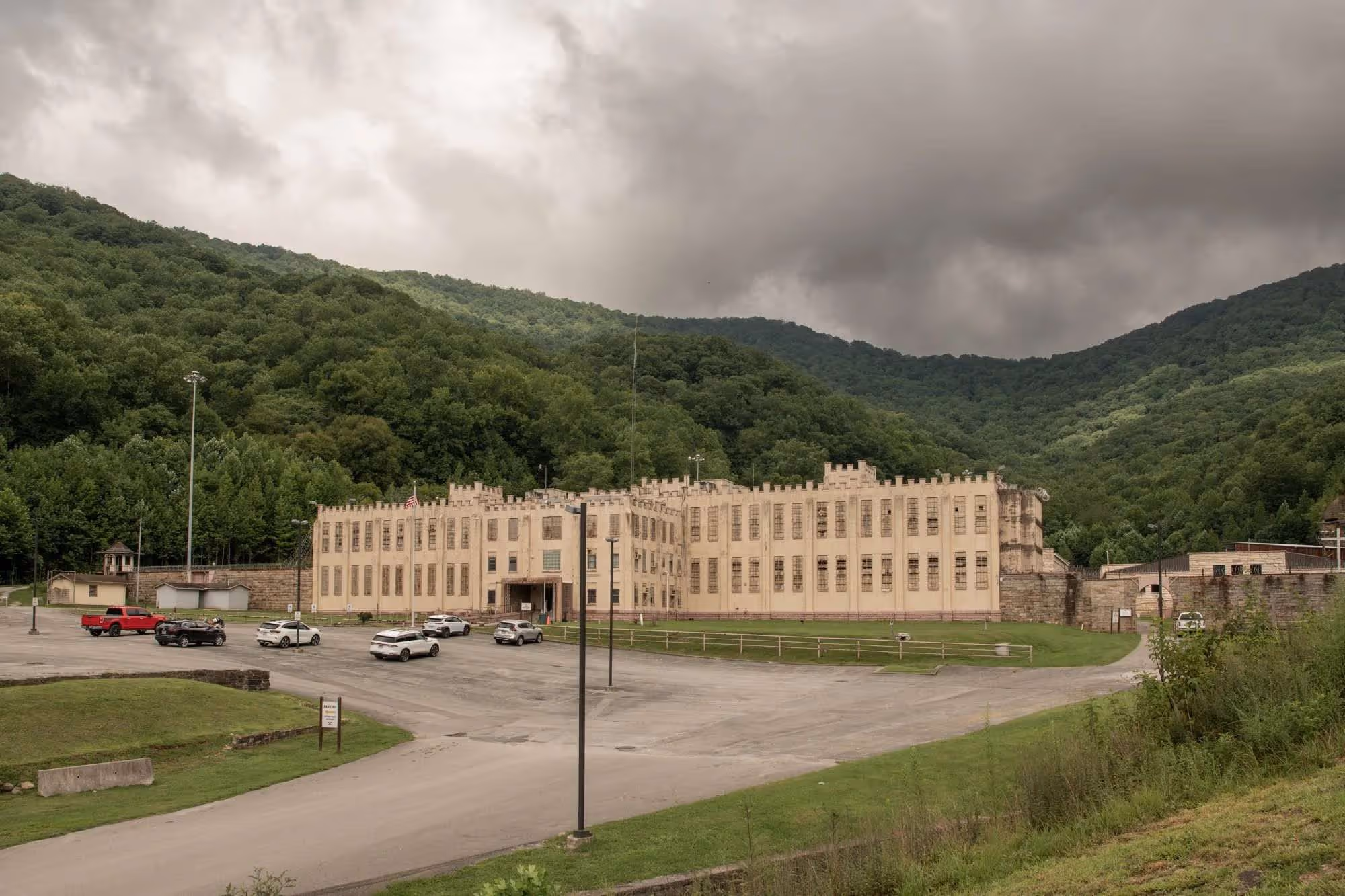 Large beige prison building with barred windows, surrounded by parking lot and green forested hills under cloudy sky.