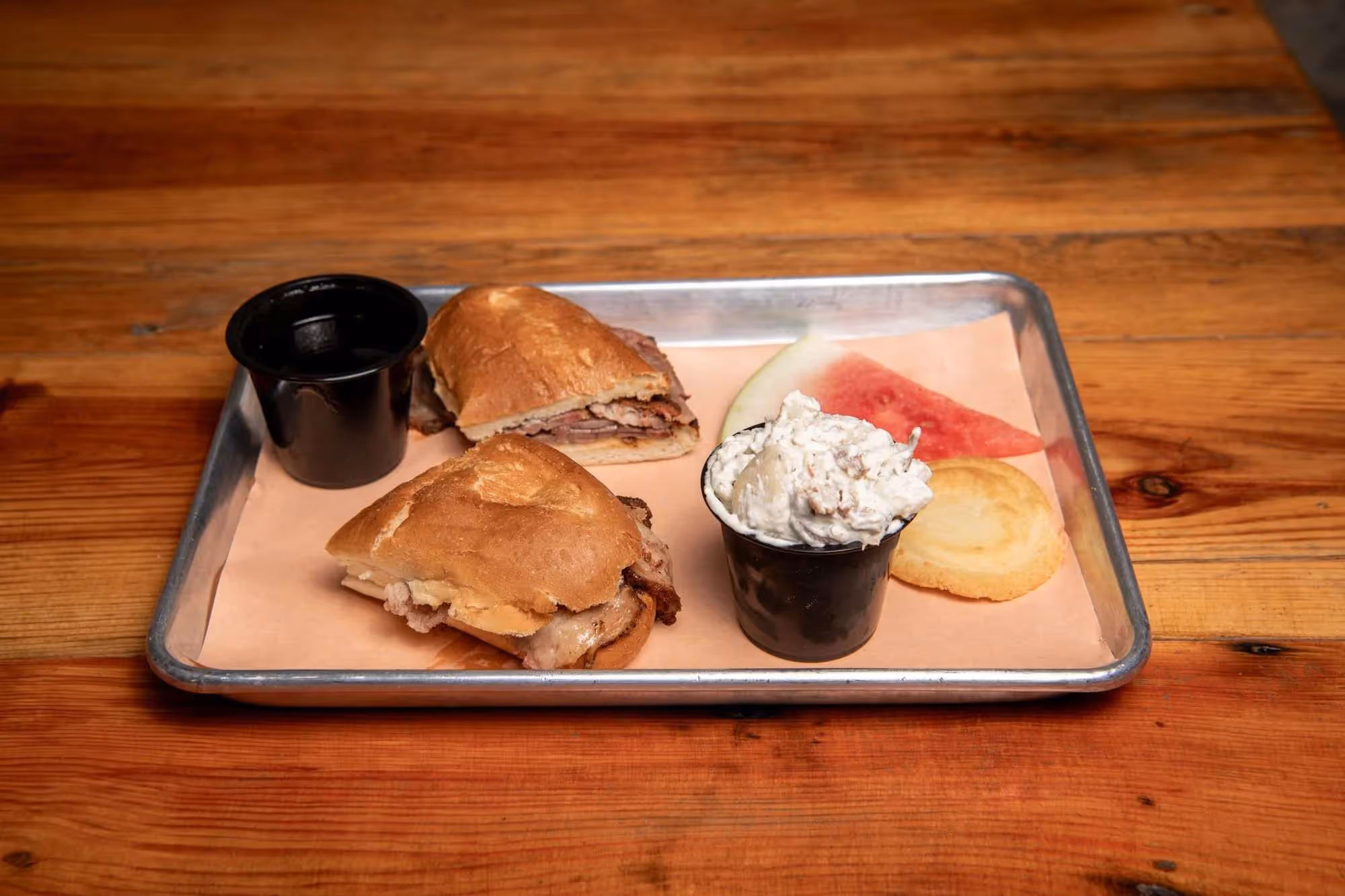 Tray with two small sandwich halves, two black cups filled with sauce and macaroni salad, a slice of watermelon, and a cookie on a wooden table.
