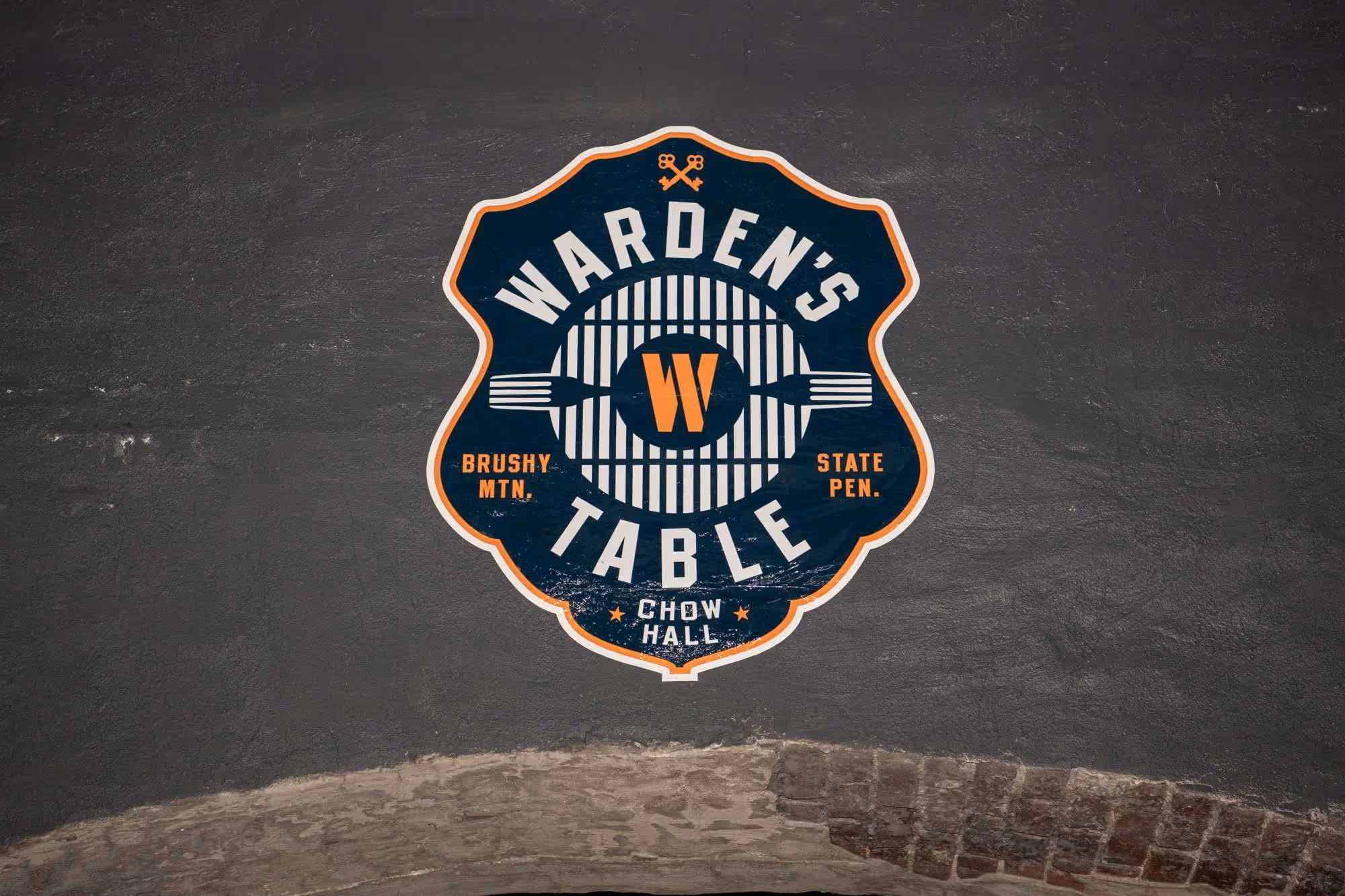 Warden's Table Chow Hall logo featuring a fork and prison bar design on a dark wall.