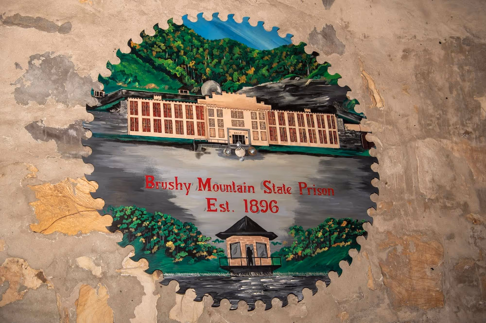 Painting of Brushy Mountain State Prison Established 1896 on a giant saw blade with surrounding green hills and a guard tower, on a worn wall.