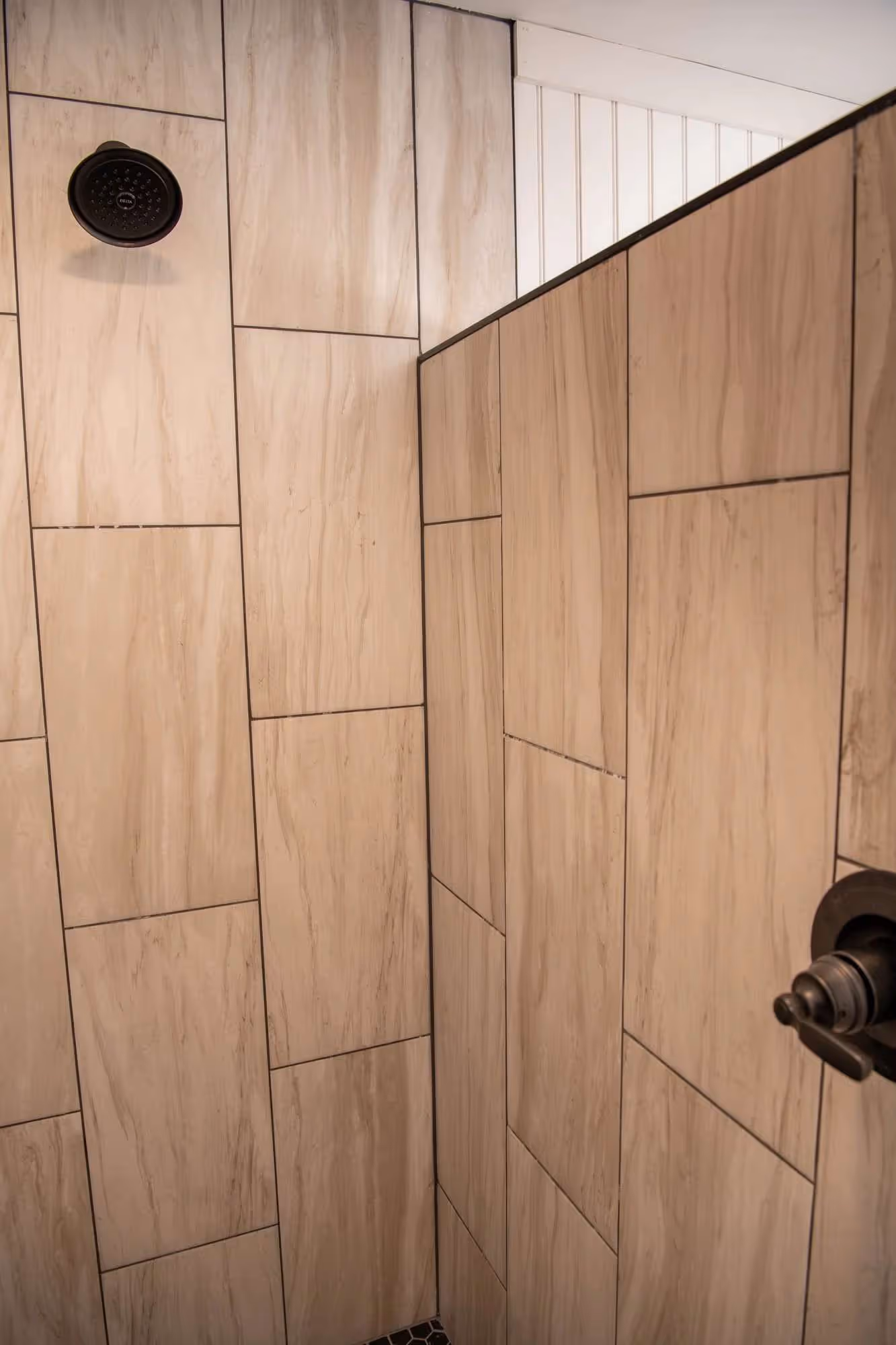 Shower interior with large beige tiles on walls, black showerhead on left wall, and black handle on right wall.