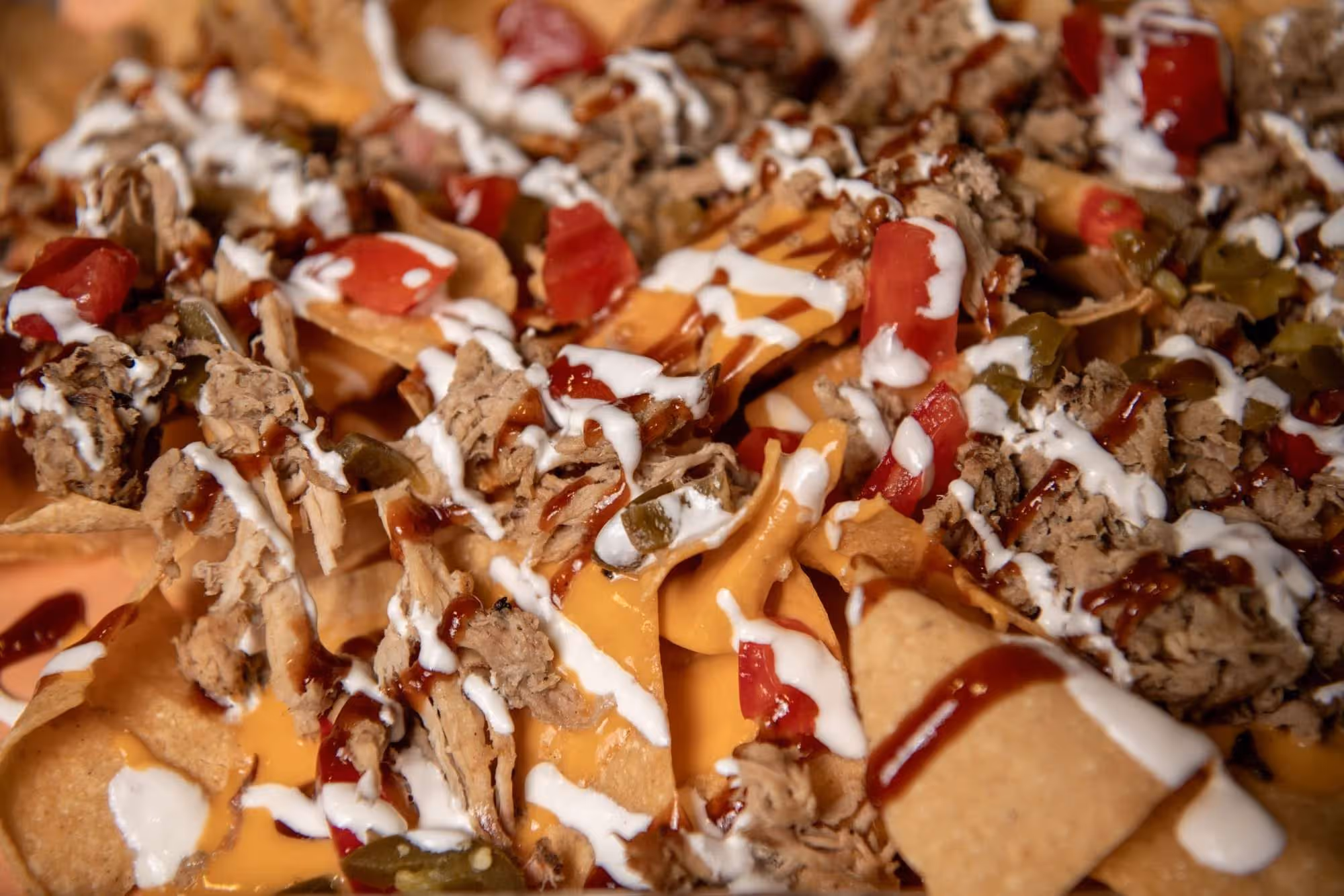 Close-up of loaded nachos topped with melted cheese, shredded meat, sliced jalapeños, diced tomatoes, and drizzled with sour cream and barbecue sauce.