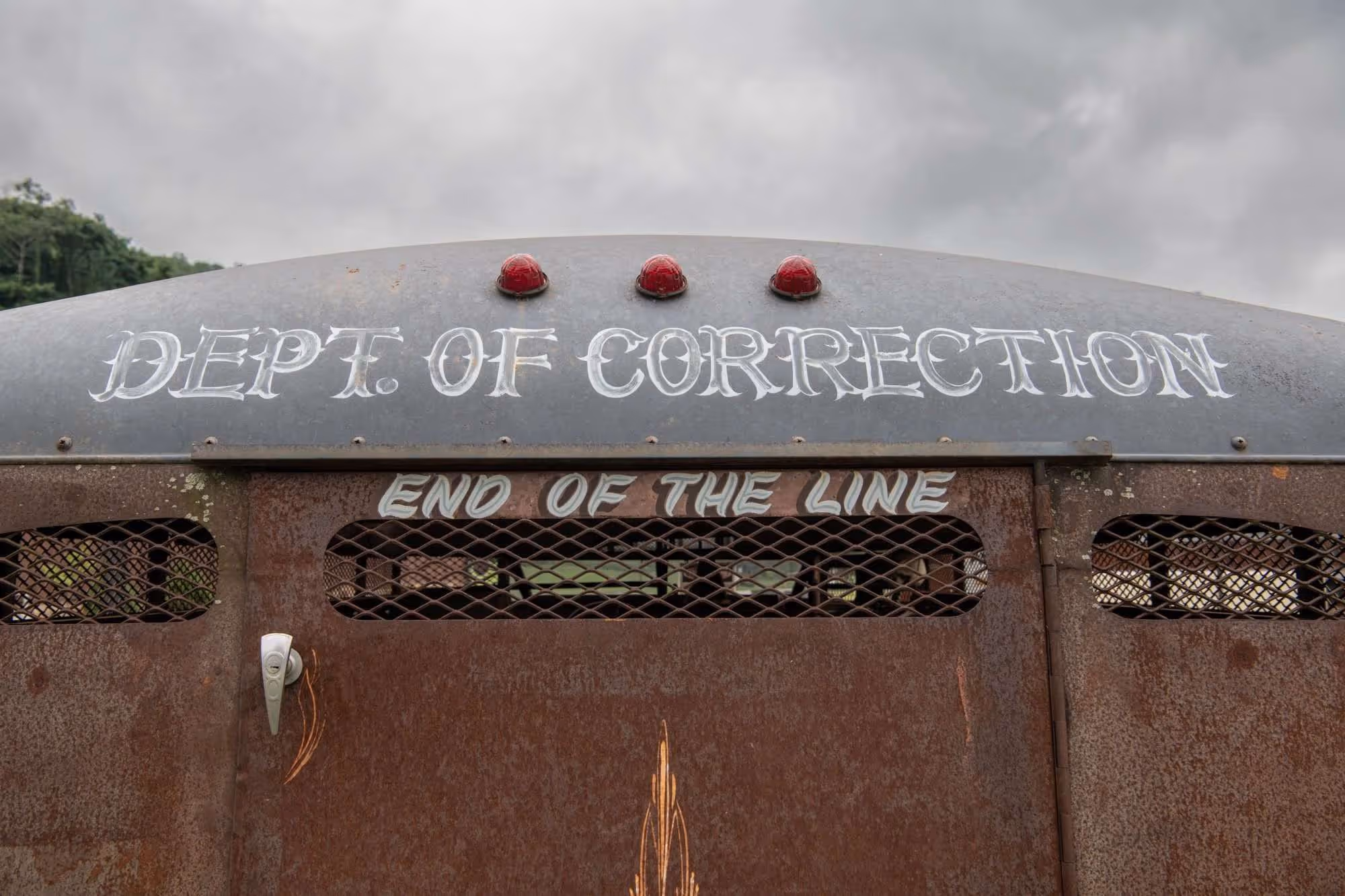 Rusty metal rear surface of a vehicle with faded white text reading 'DEPT. OF CORRECTION' and 'END OF THE LINE' under a cloudy sky.