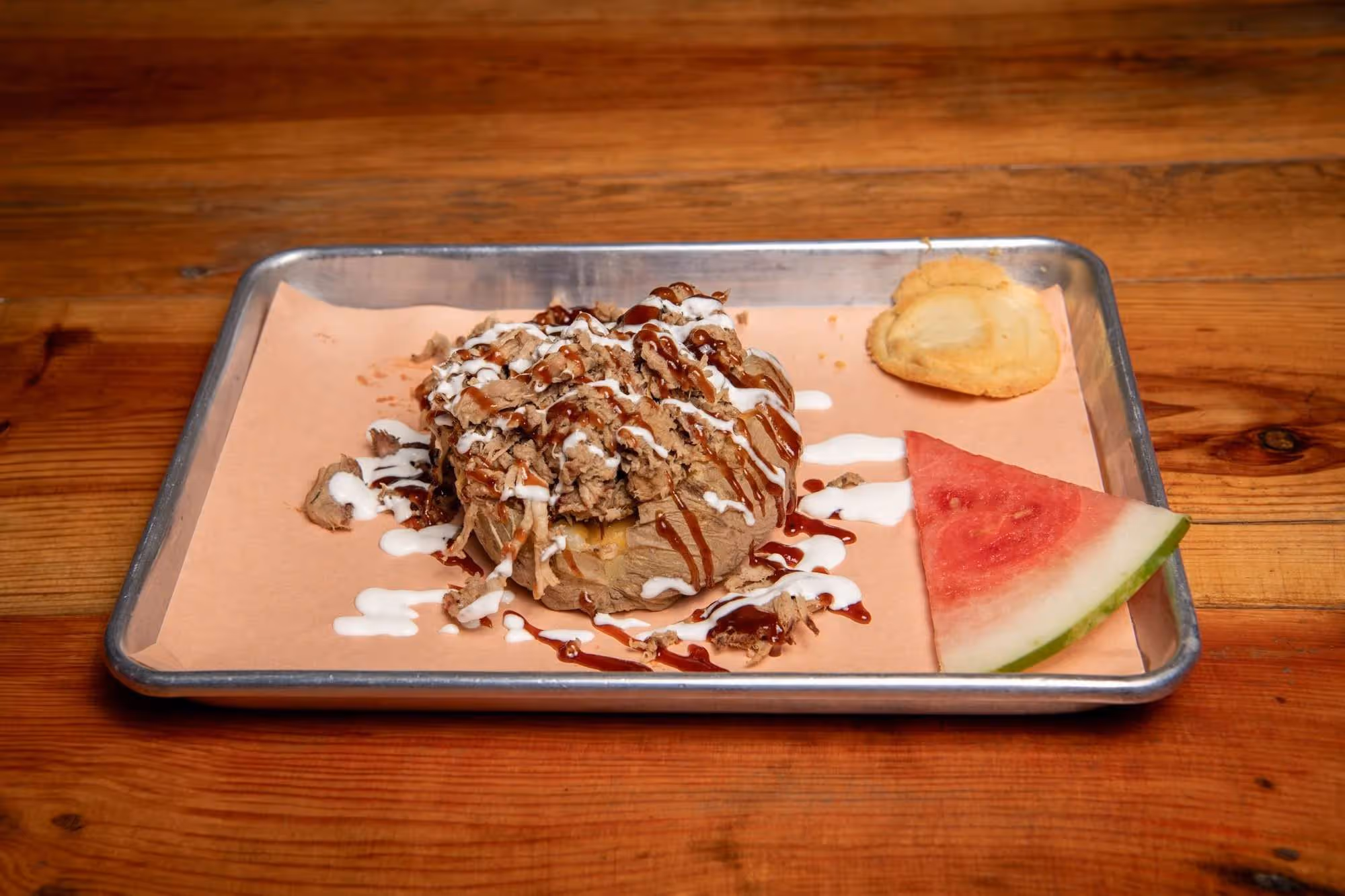 Barbecue baked potato on a tray with barbecue and sour cream, served with a slice of watermelon and a cookie.