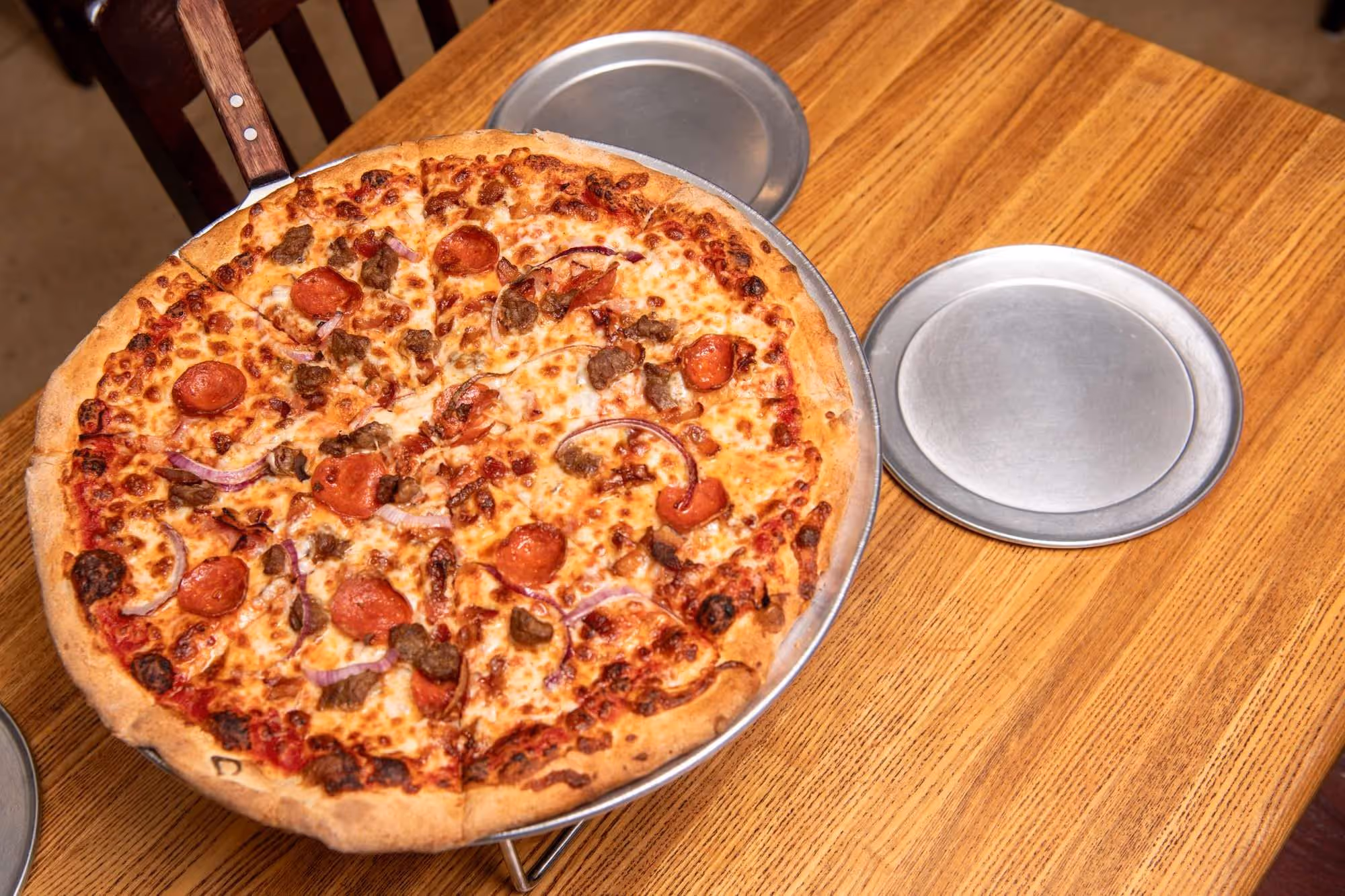 Pepperoni and beef pizza with red onion slices on a metal tray on a wooden table next to empty metal plates.
