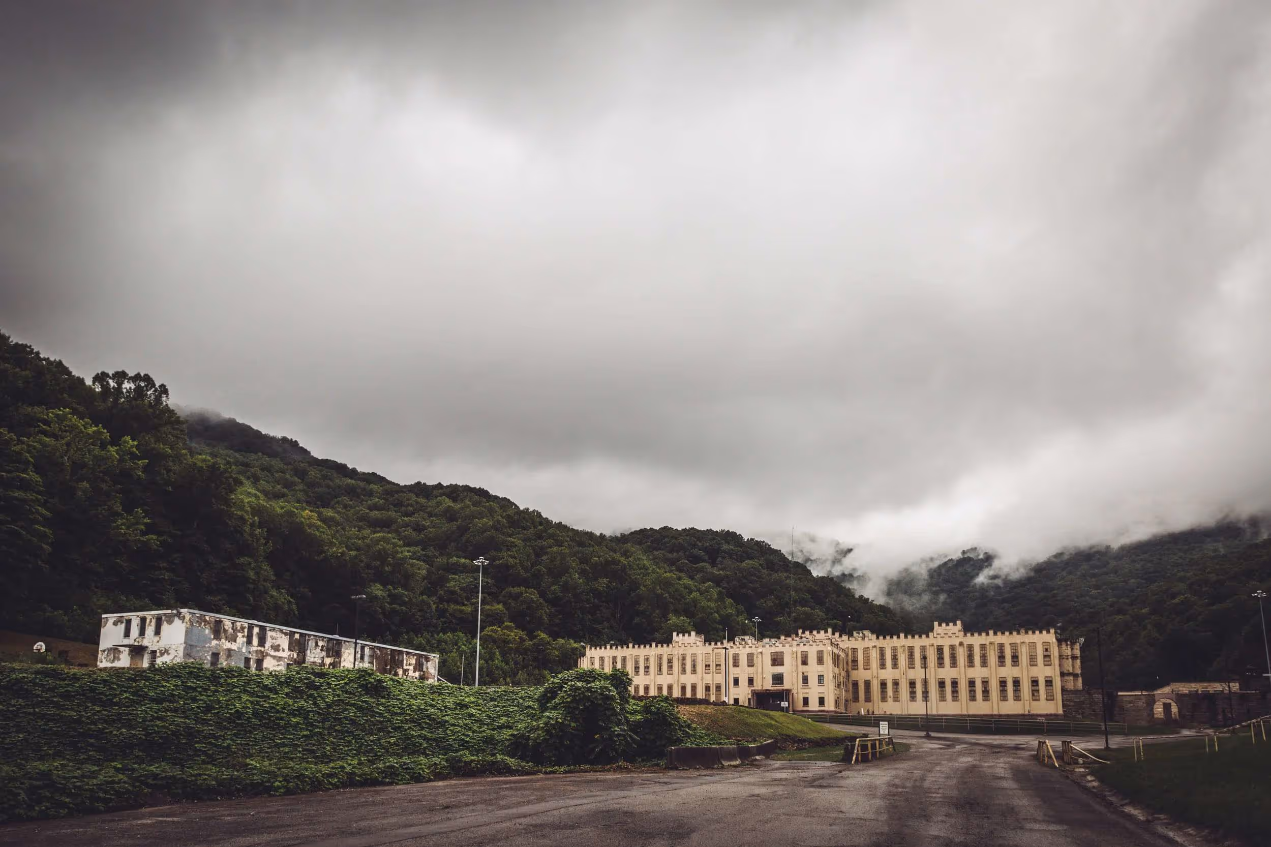 Large historic prison building set against forested hills under a cloudy sky with mist in the background.