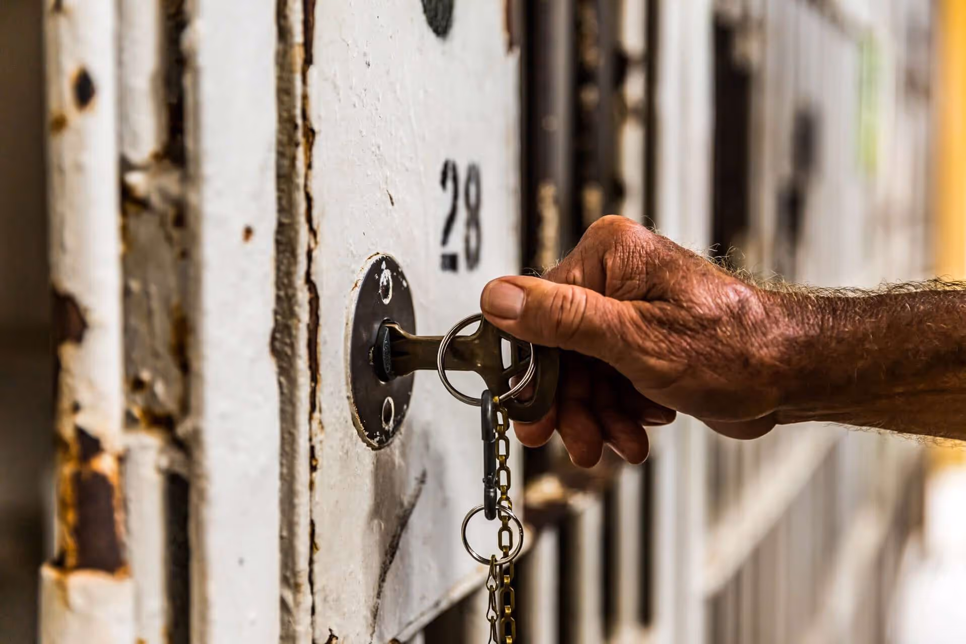 A hand inserting a key into the lock of a weathered white metal prison cell door numbered 28.