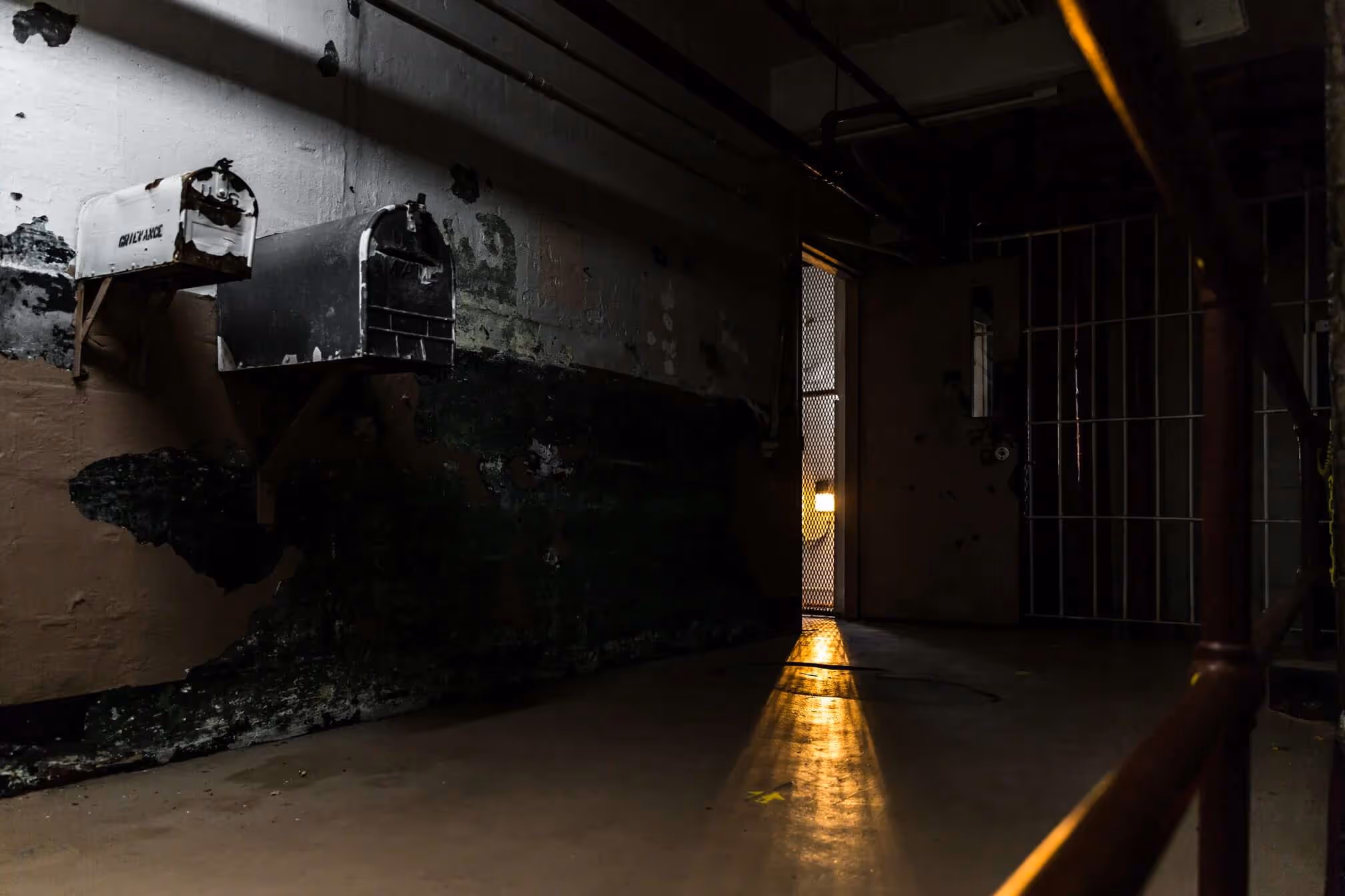 Dimly lit, aged prison cell block corridor with two old mailboxes on the wall and a door ajar casting a warm light on the floor.