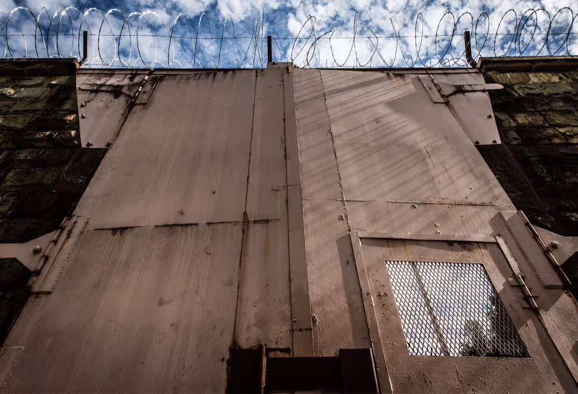 Close-up view of a tall metal prison gate with barbed wire on top against a cloudy sky.